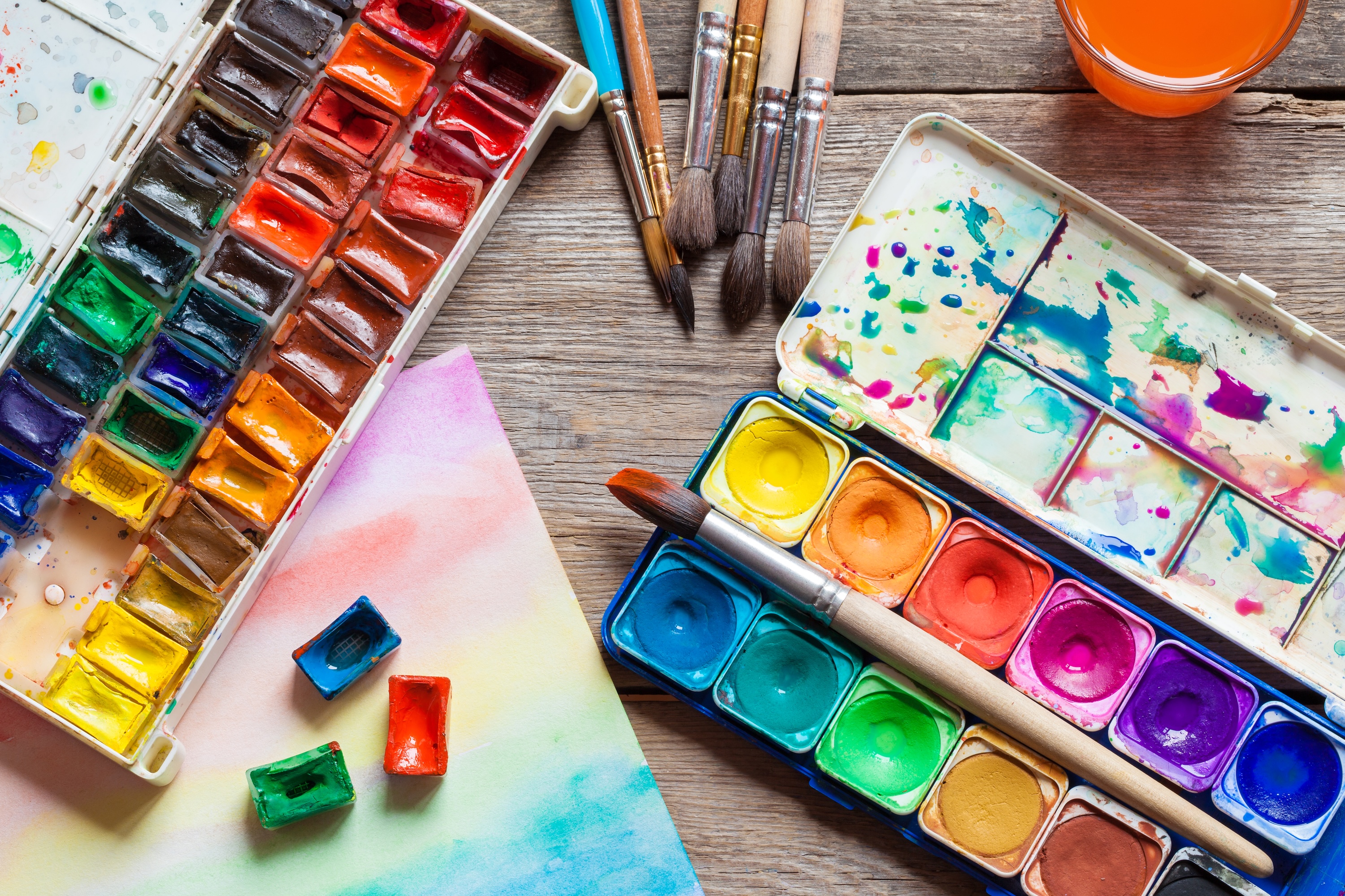 A close up of watercolour and oil based paints along with some paint brushes, some paint water used to clean brushes and a paper started with watercolour
