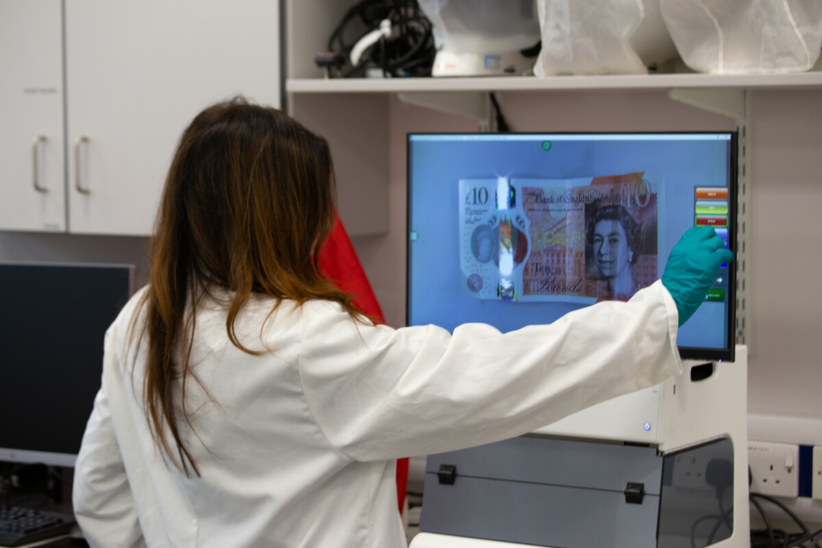 A Forensic Sciences student dressed in a white laboratory coat uses the Video Spectral Comparator to examine a £10 bank note in a USW forensic laboratory