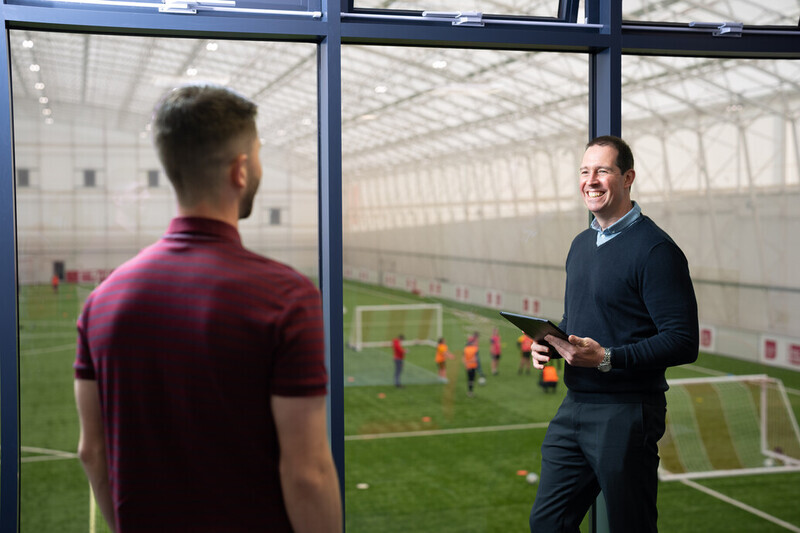 Two individuals talking and smiling overlooking an indoor football pitch.