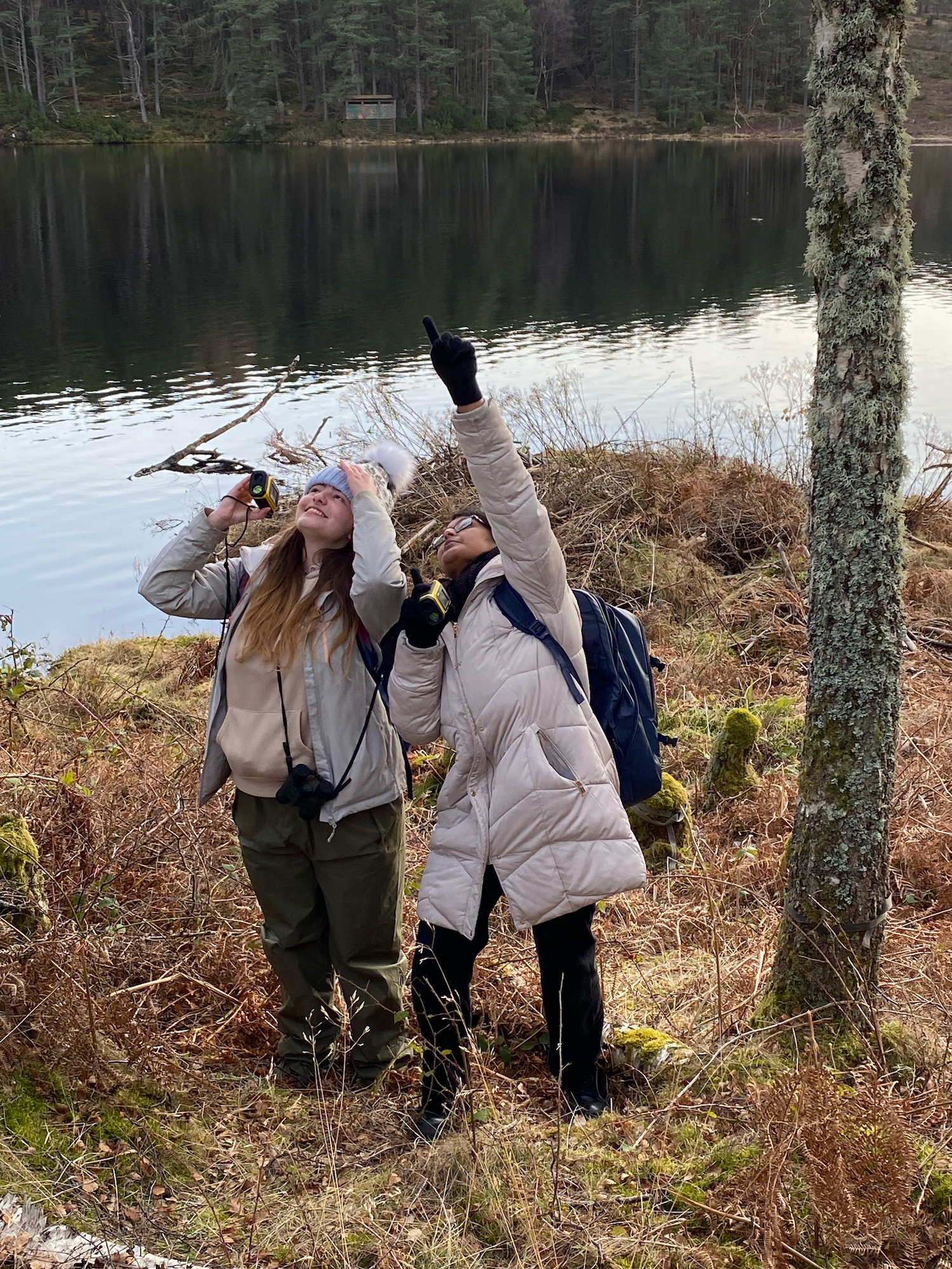 Two students wearing warm-looking coats, waterproof trousers, winter hats and gloves point at the sky and smile while using recording equipment on a mossy bank near a lake with trees