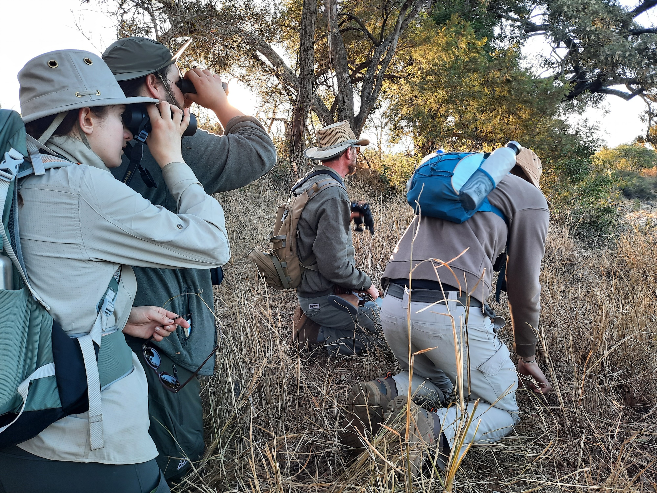 A group of international wildlife students crouch in long grass while looking through binoculars into the distance while on a field trip in South Africa