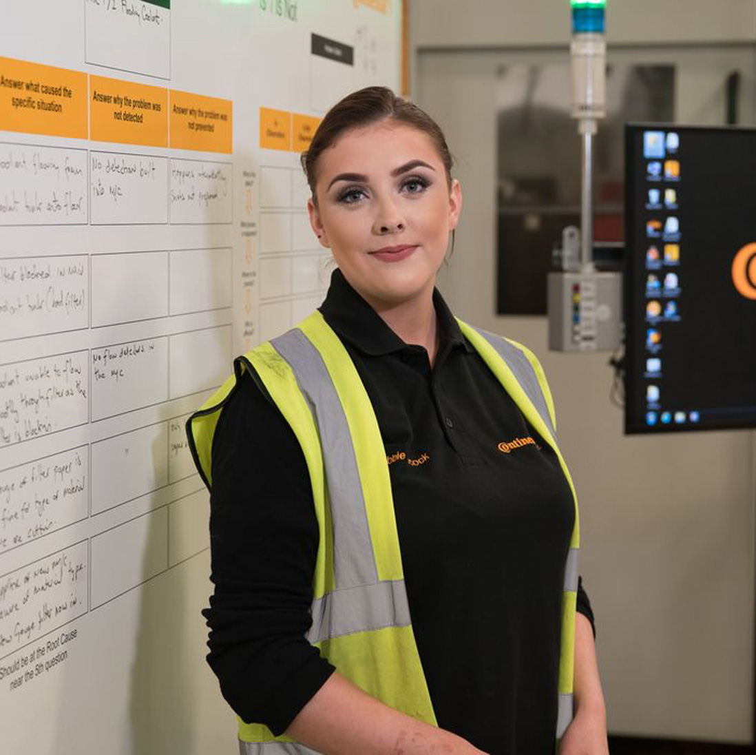 A logistics, procurement and supply chain management student smiles at the camera while wearing a yellow high visibility jacket in front of a whiteboard in an office
