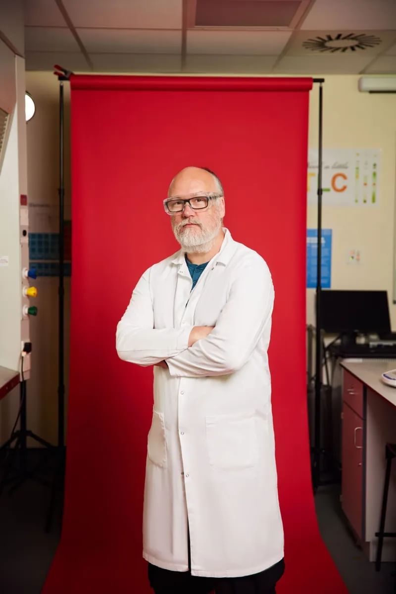 An academic wearing a white laboratory coat looks at the camera with their arms folded in front of a red backdrop inside a USW laboratory at Glyntaff campus