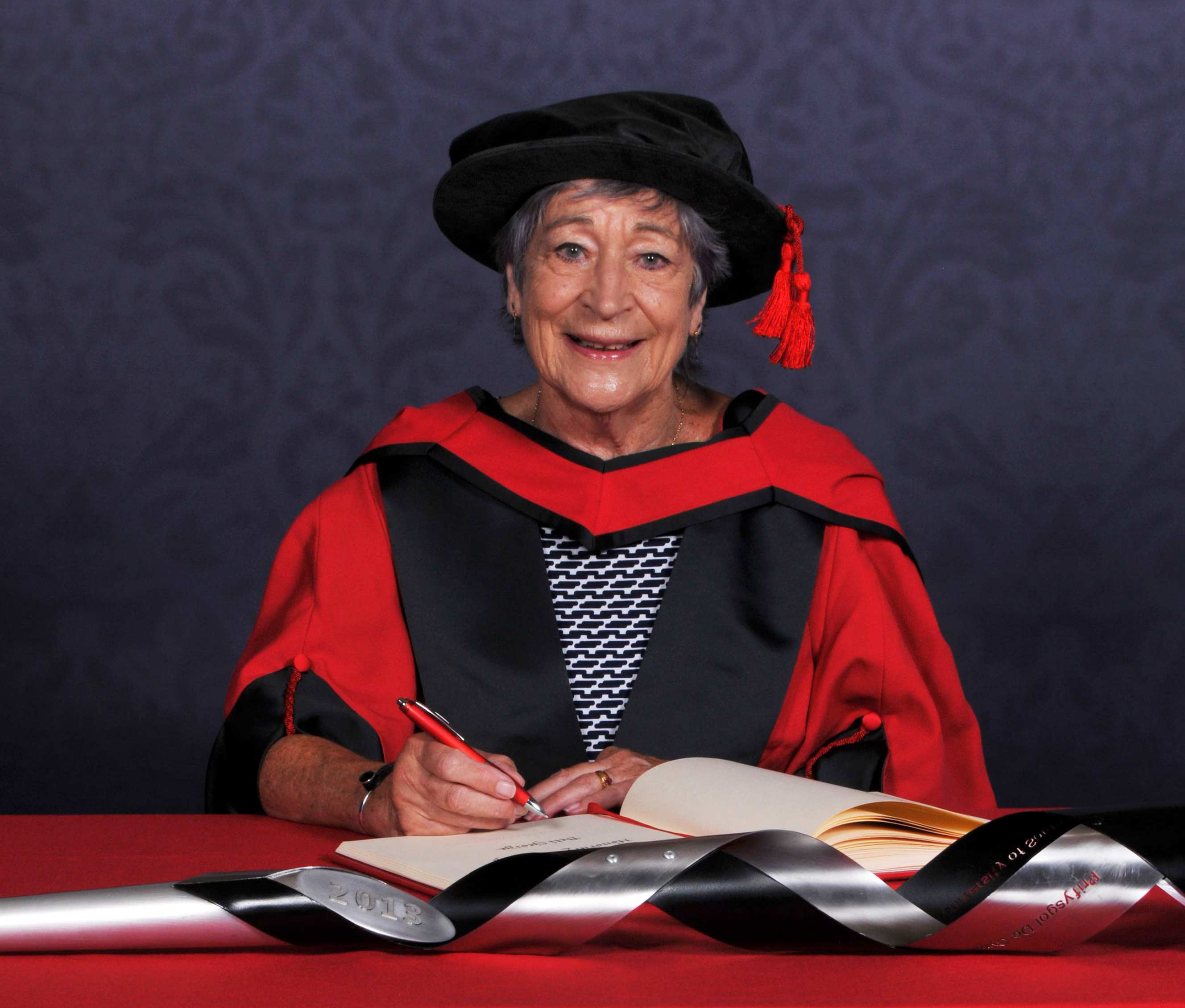 Beti George signs a book on a red desk while wearing an honorary graduate's cap and gown in front of a blue backdrop
