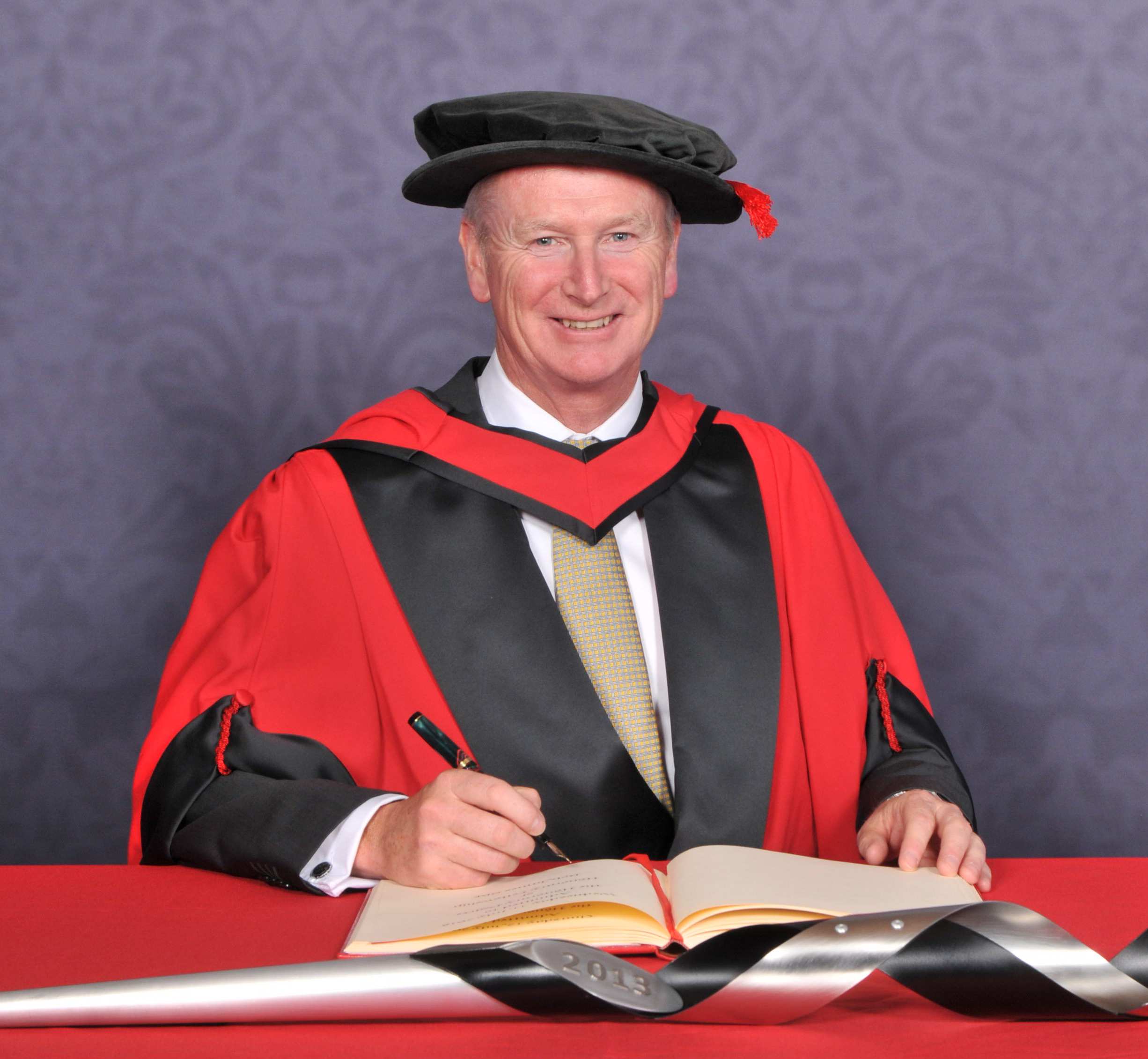 Bob James OBE smiling while signing a large book on a red desk and wearing an honorary graduate cap and robe in front of a blue backdrop