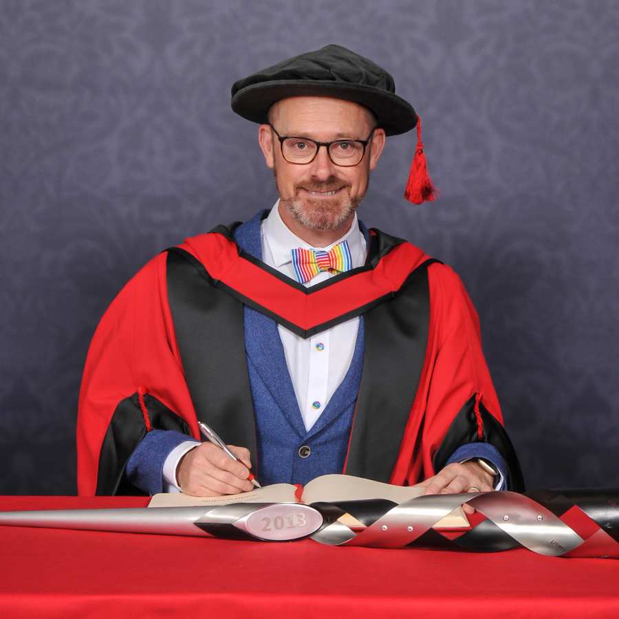Honorary fellow Andrew White looks at the camera as they write in a large book on a red desk while wearing a PhD cap and gown