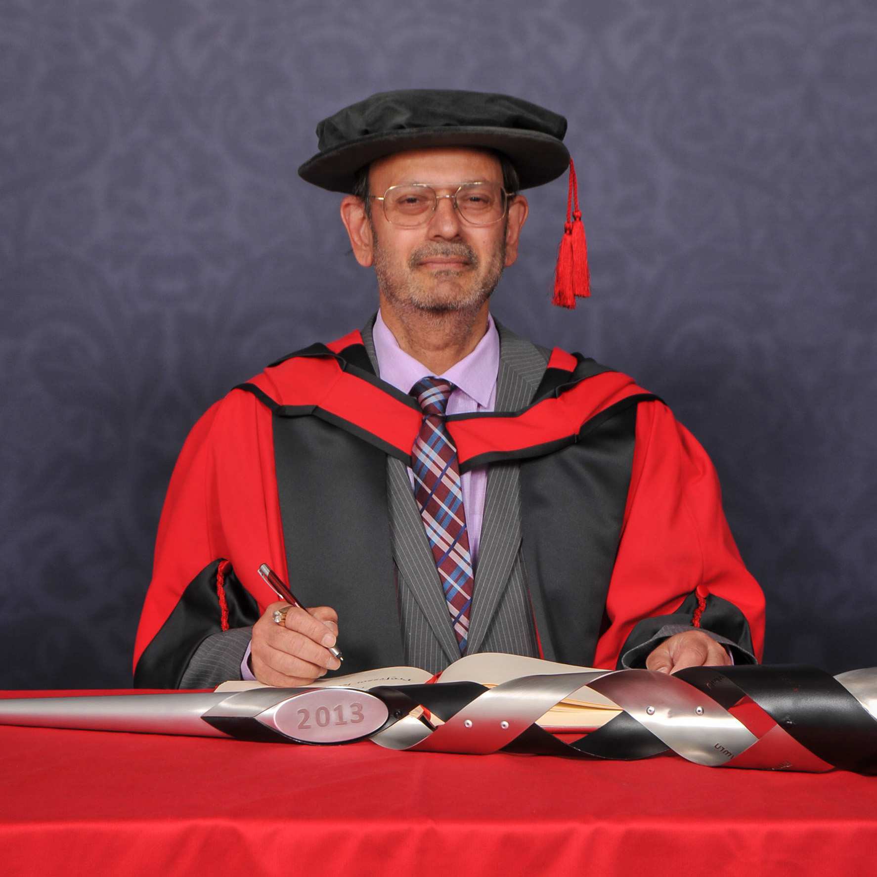 Honorary fellow Keshav Singhal looks at the camera as they write in a large book on a red desk while wearing a PhD cap and gown