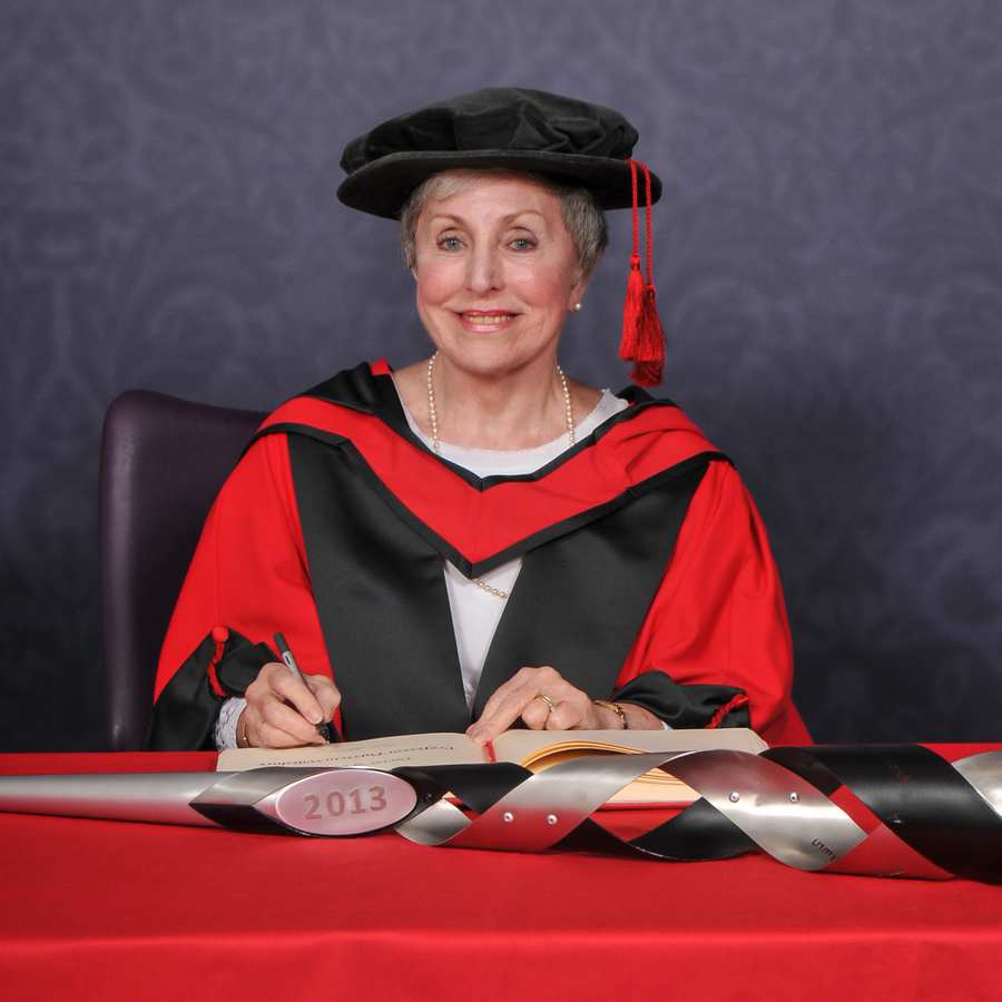 Honorary fellow Patricia Wiltshire smiles at the camera as they write in a large book on a red desk while wearing a PhD cap and gown