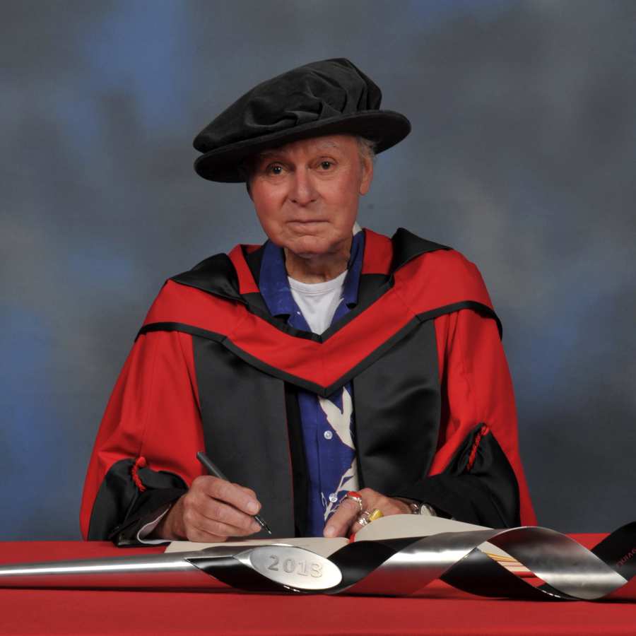 John Selway writes in a large book next to a ceremonial mace on a red desk while wearing a doctorate's cap and gown in front of a blue backdrop.