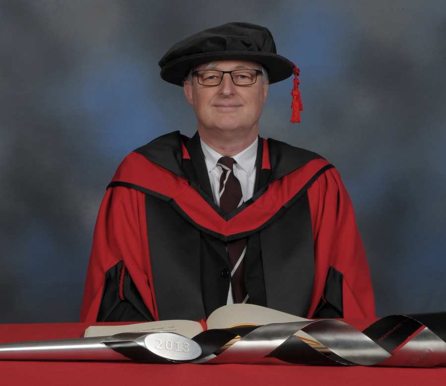 Rhobert Lewis writes in a large book next to a ceremonial mace on a red desk while wearing a doctorate's cap and gown in front of a blue backdrop.