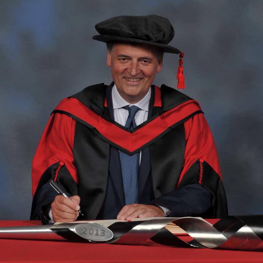 Steven Moffat writes in a large book next to a ceremonial mace on a red desk while wearing a doctorate's cap and gown in front of a blue backdrop.