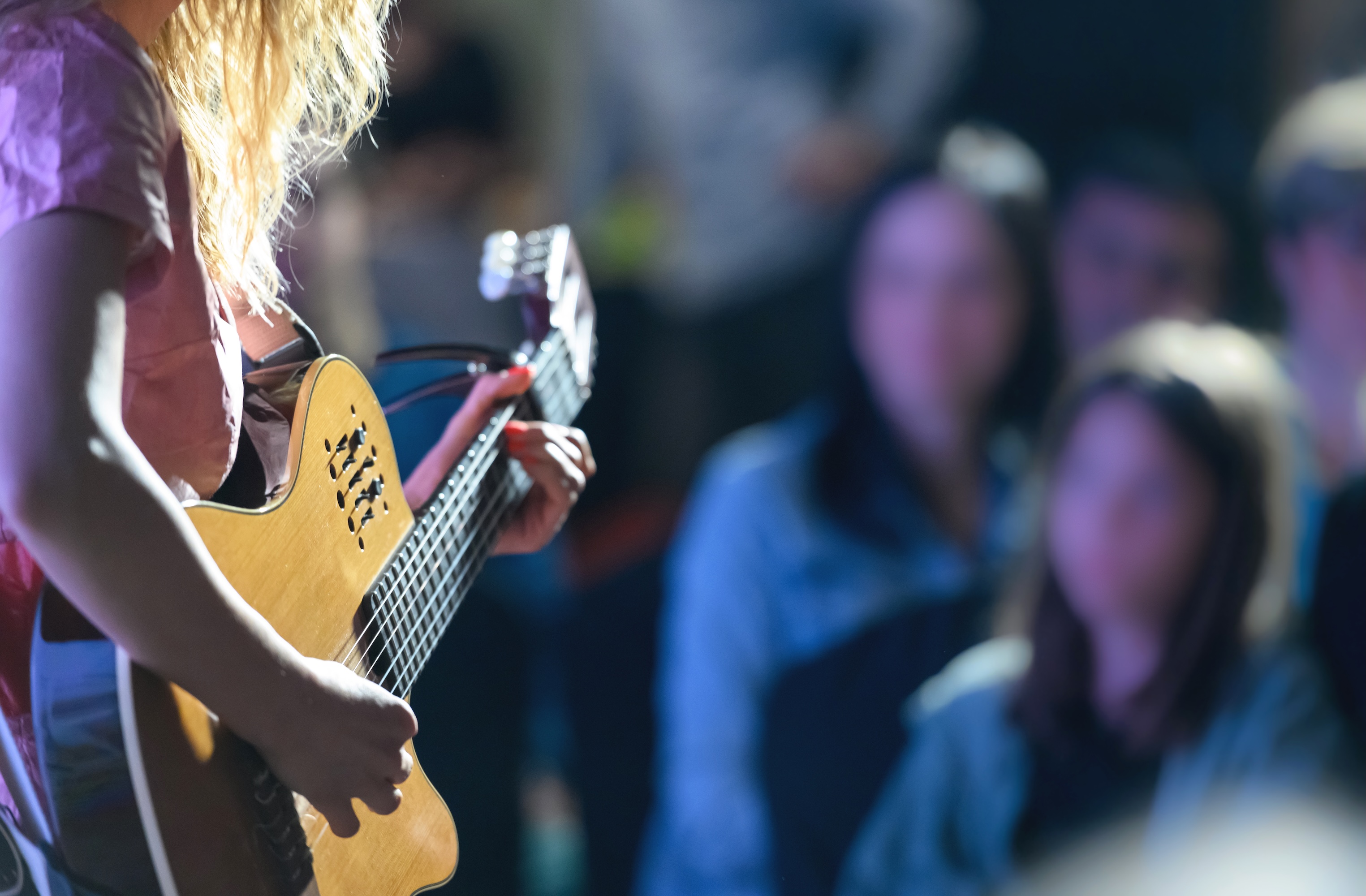 The side of musician with long blond hair playing a yellow semi-acoustic guitar on a stage with a blurred out audience in the background watching