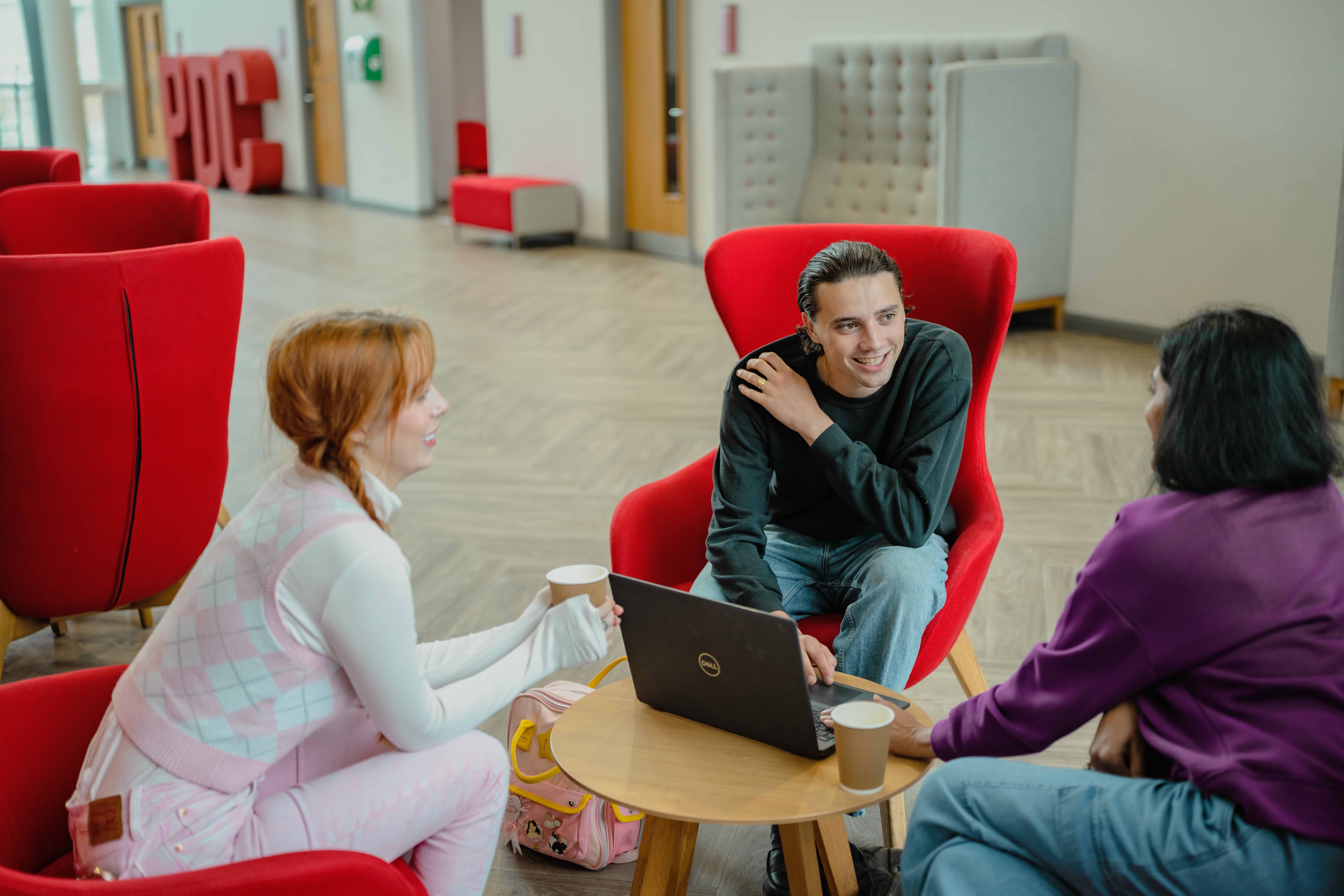 Students sat in a communal area on campus, working on a laptop and smiling.