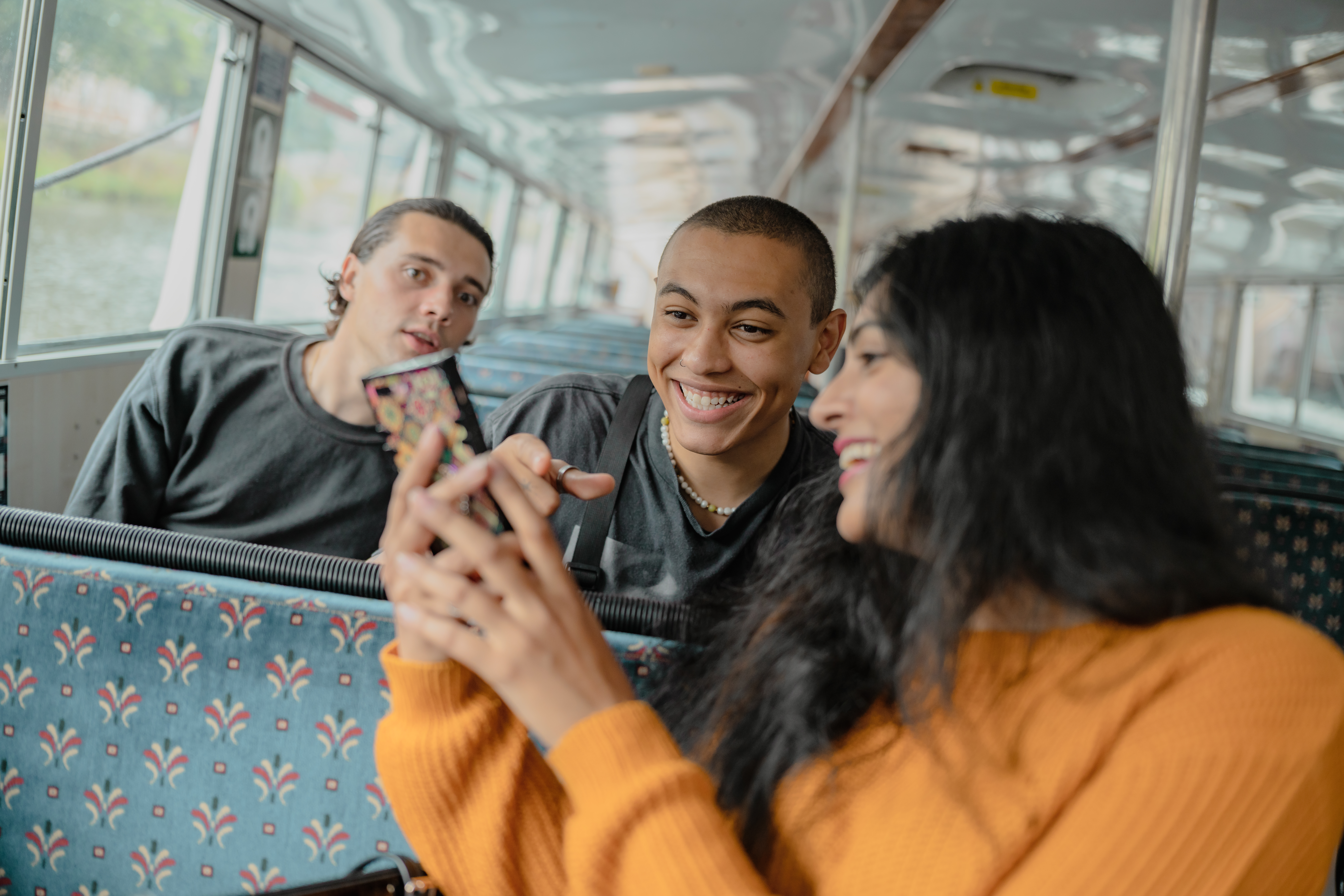 Three students are sat together on a bus. One of them is turning around to show the other two her mobile phone and they are laughing.