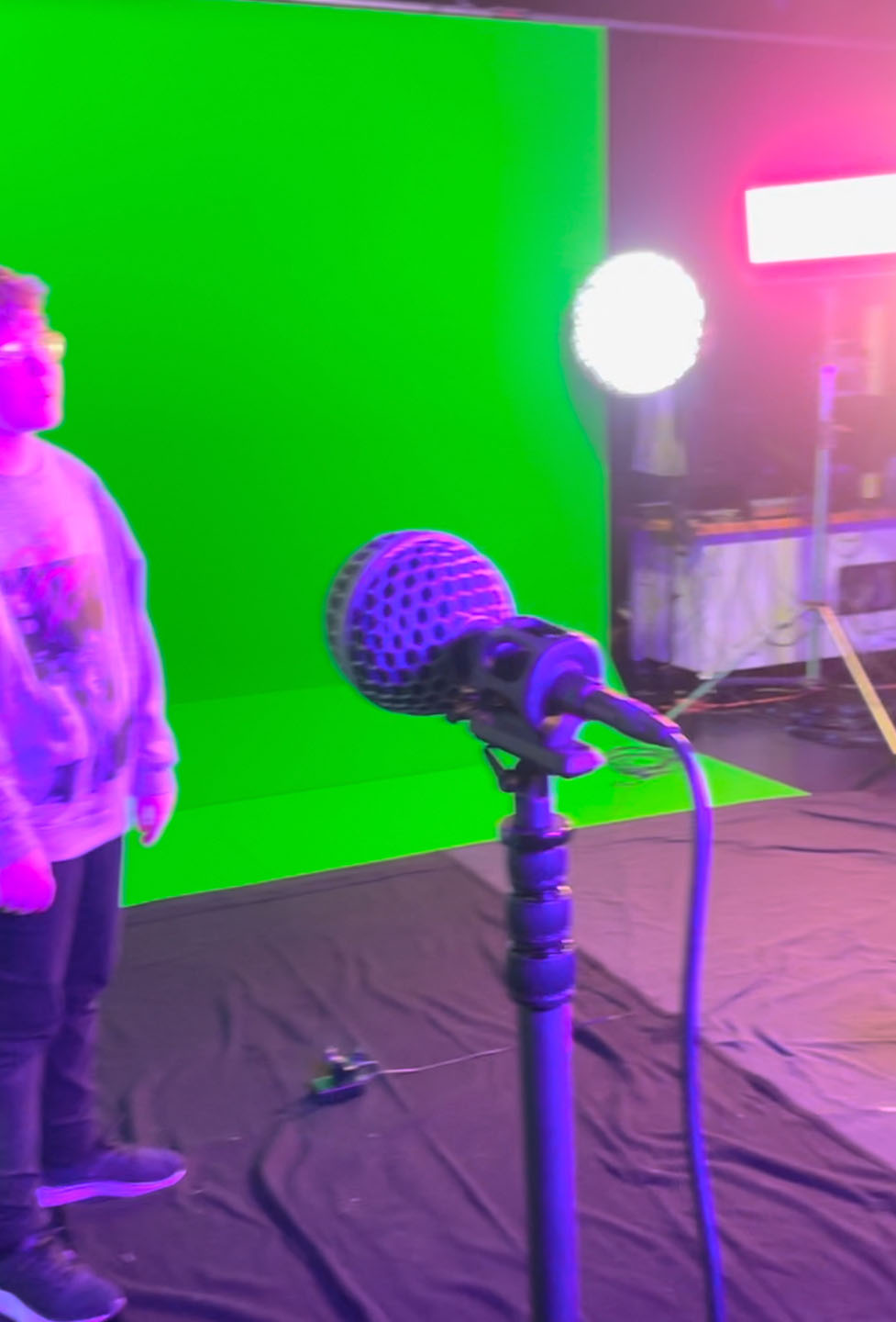 A microphone on a stand. There is a person stood near a green screen in the background.