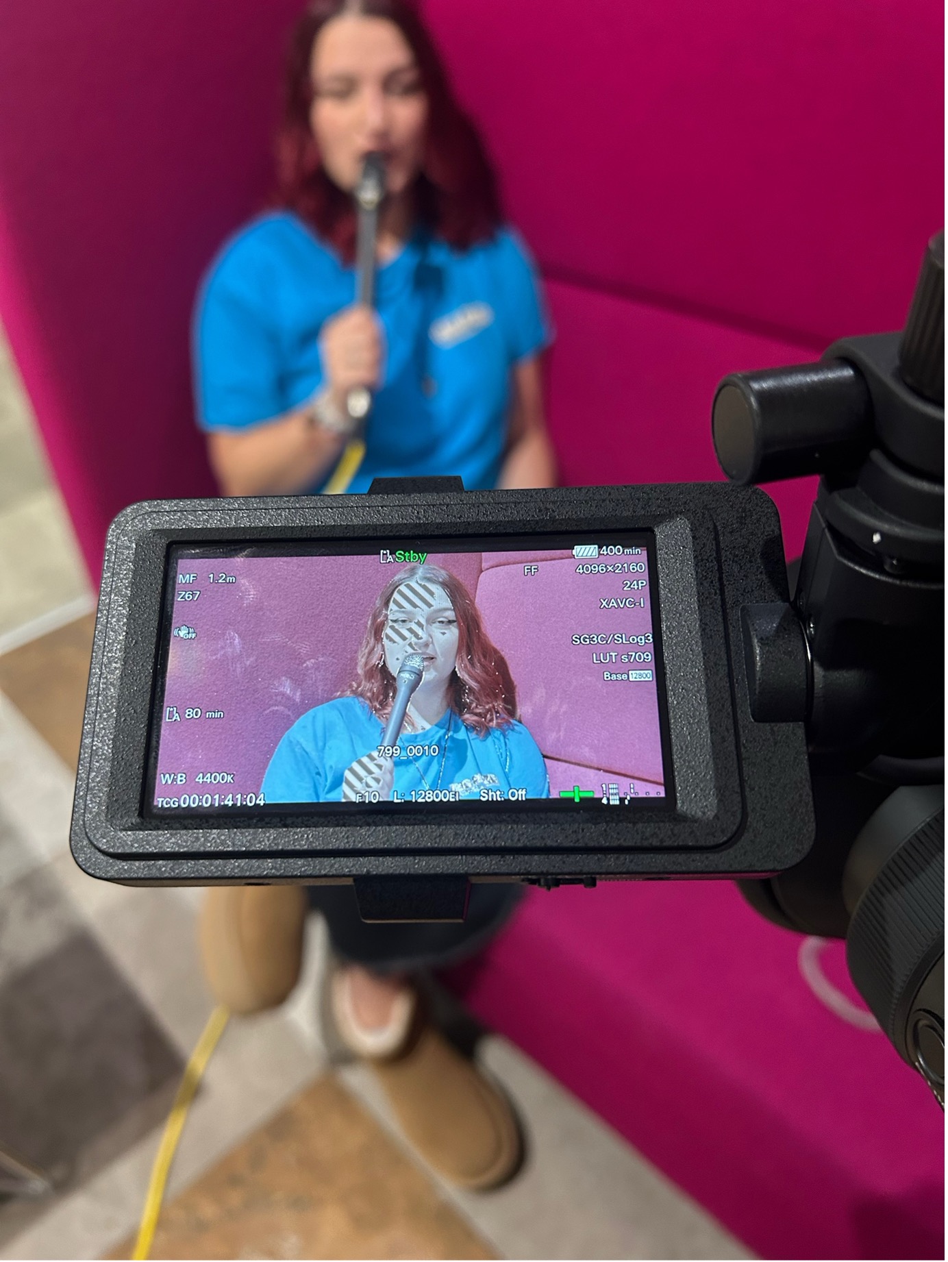 A camera screen focused in on a young woman holding a microphone to her mouth.