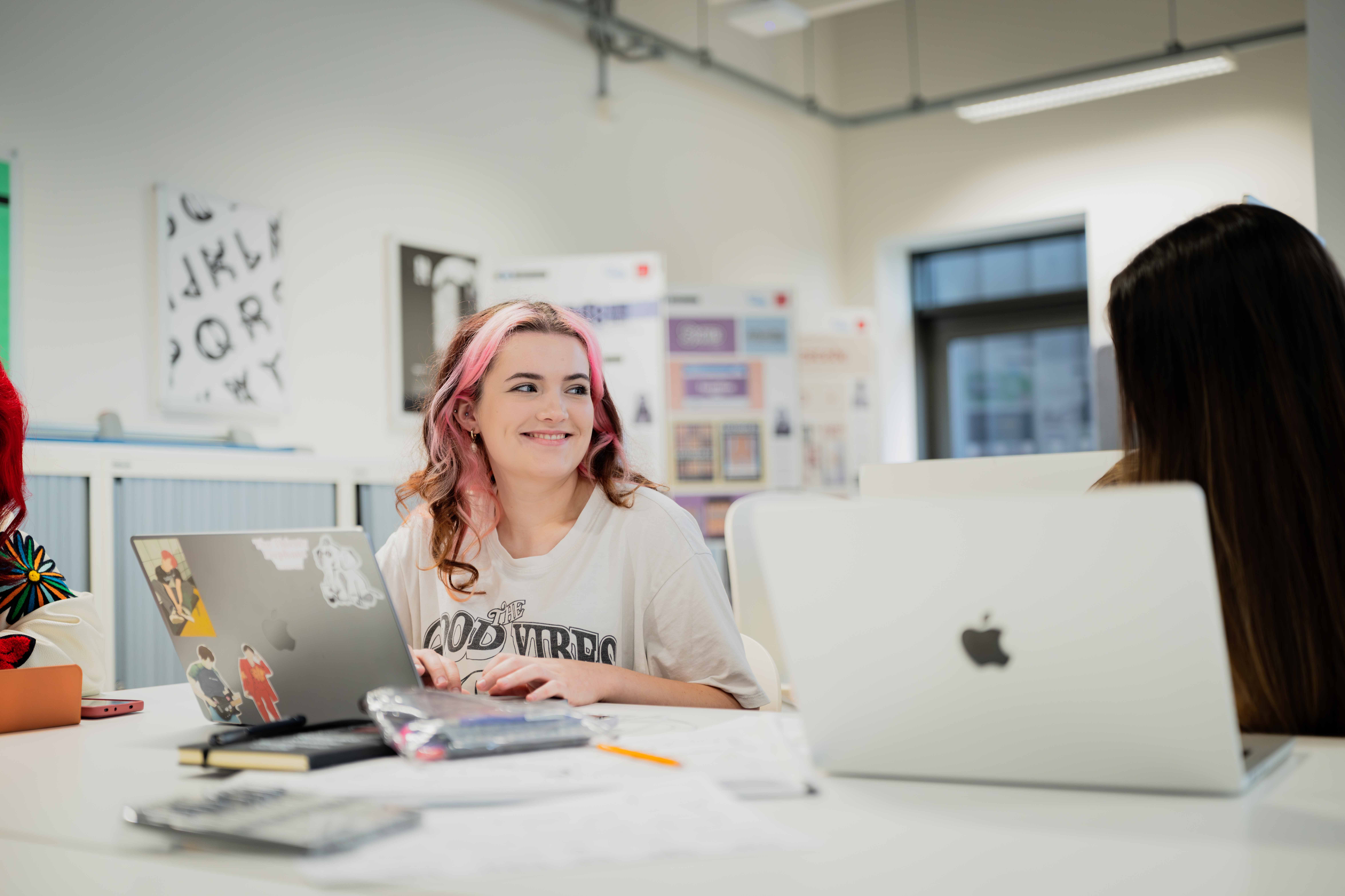 A student with pink and brown hair is sitting in a studio, working on a laptop and smiling.