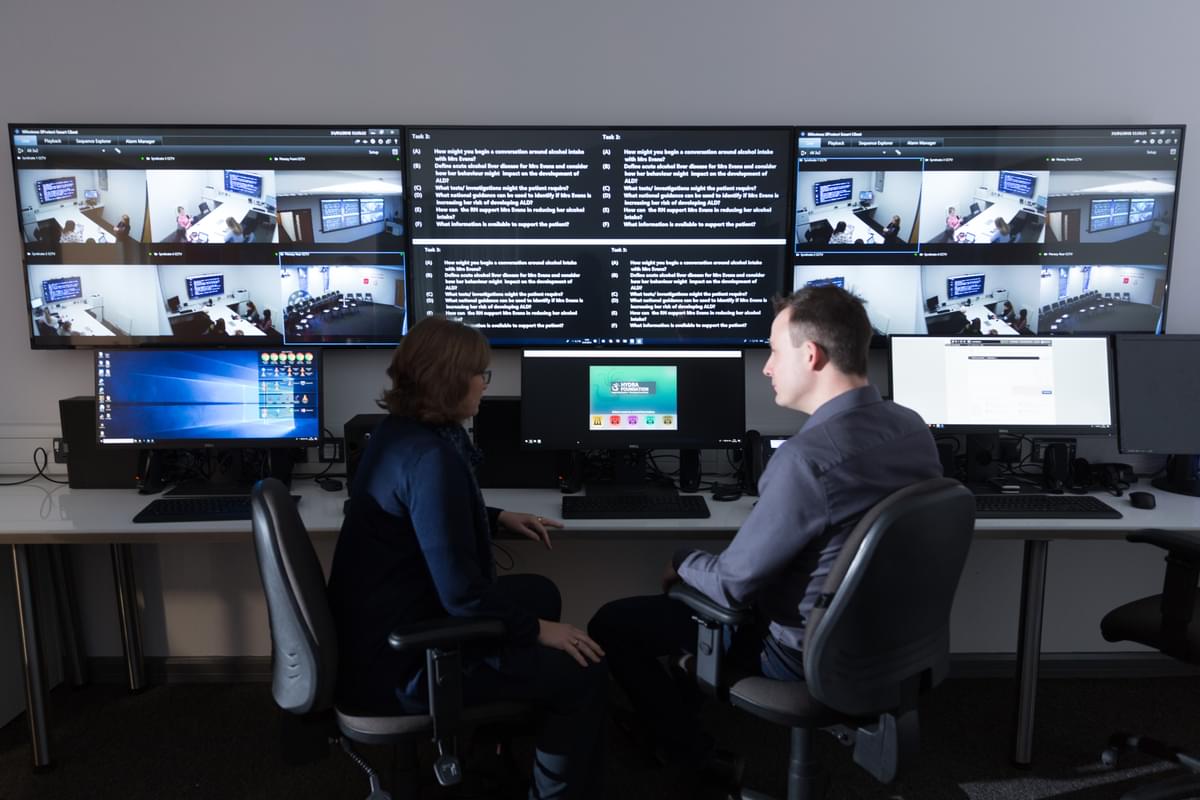Two policing lecturers sat discussing in front of the screens in the hydra simulation centre on glyntaff campus