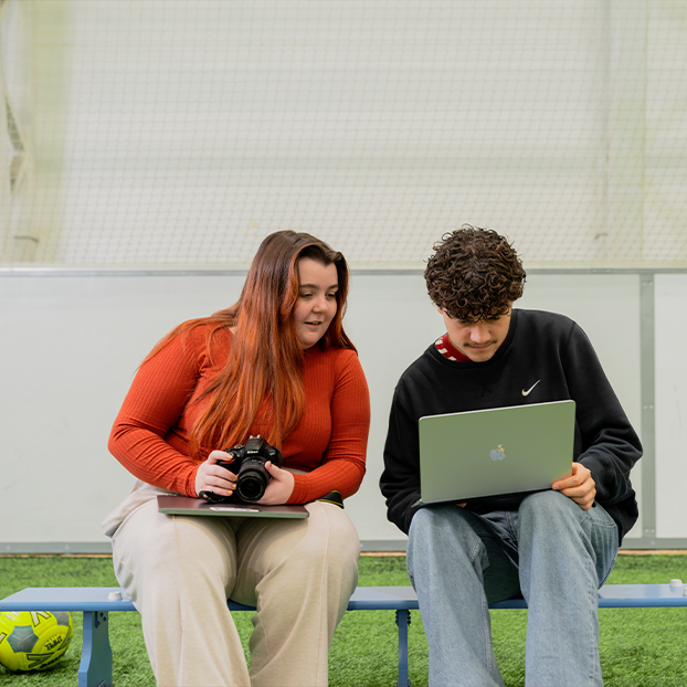 Two Sports Journalism students are sitting on a bench at the side of a football pitch and working together. One is holding a camera and one is holding a laptop.