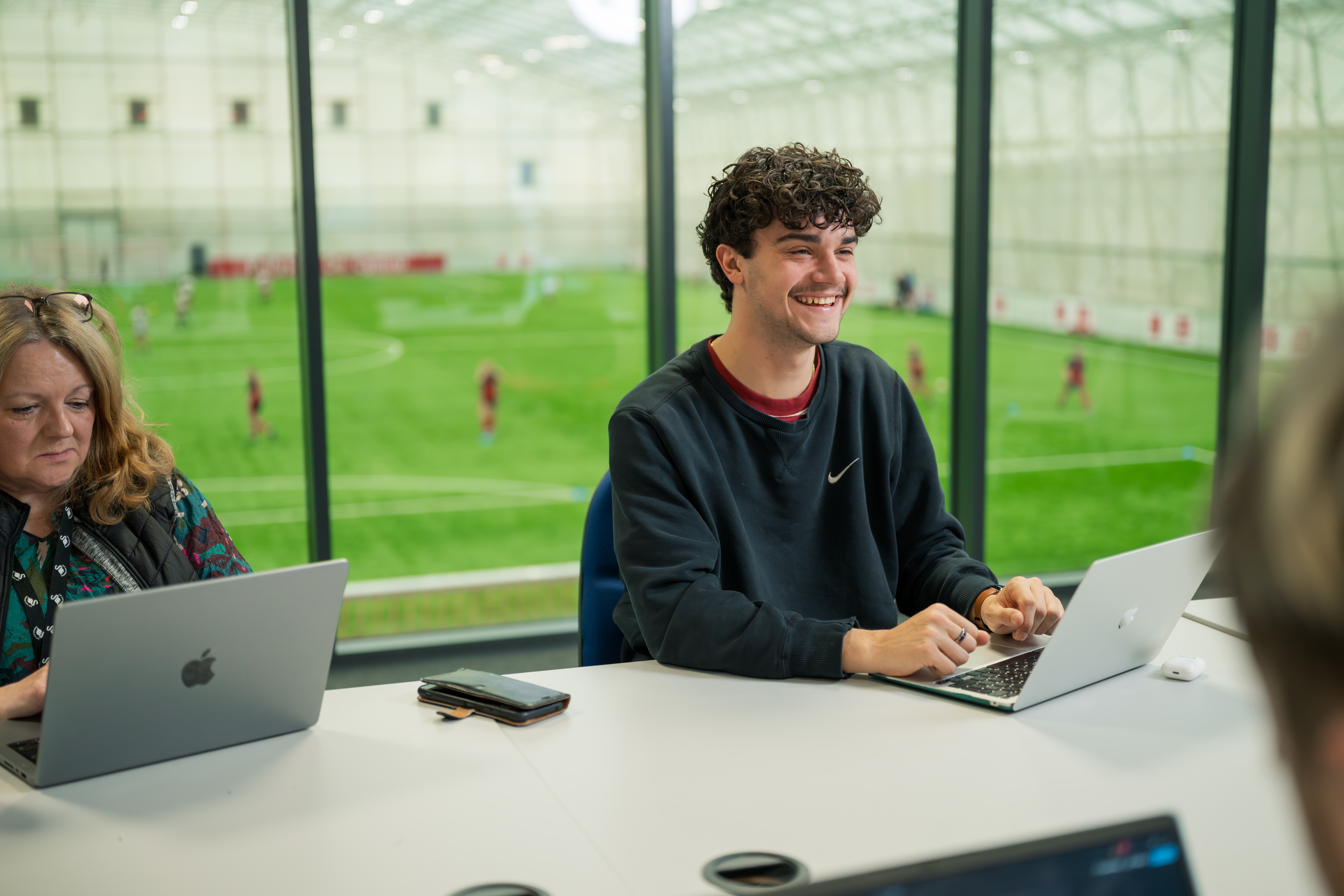 Sports Journalism student, Freddie, is sitting in a classroom with his laptop. There is a view of a football pitch through the glass windows behind him.