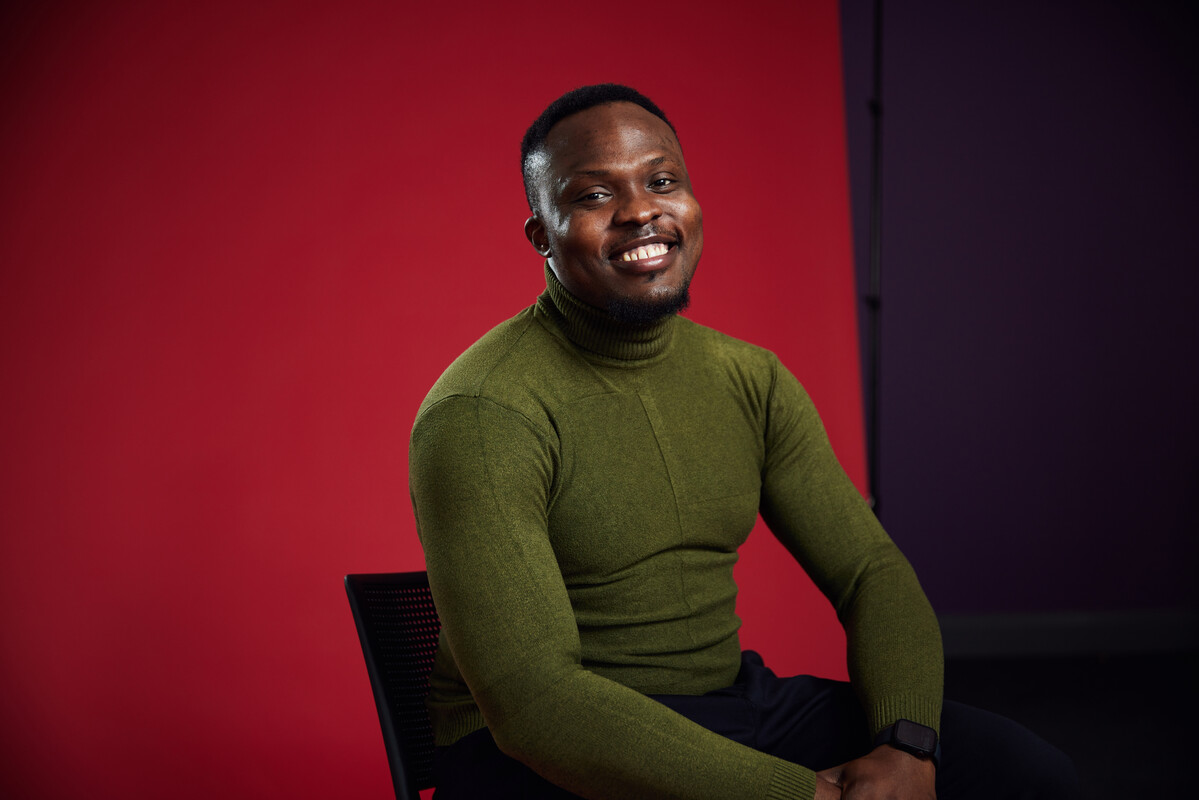 Student chidiebere Ejiogu leans on their lap and smiles at the camera while sat in front of a red backdrop