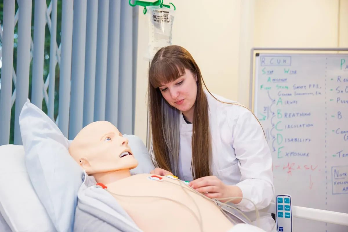 A student in scrubs works on a medical dummy in a clinical simulation center, wearing a white lab coat.