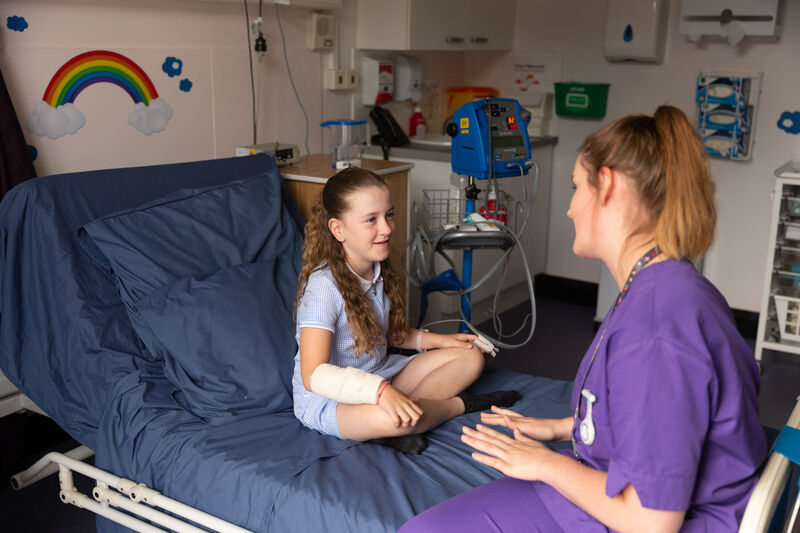 A nurse and a child with a arm cast sitting on a hospital bed in coversation