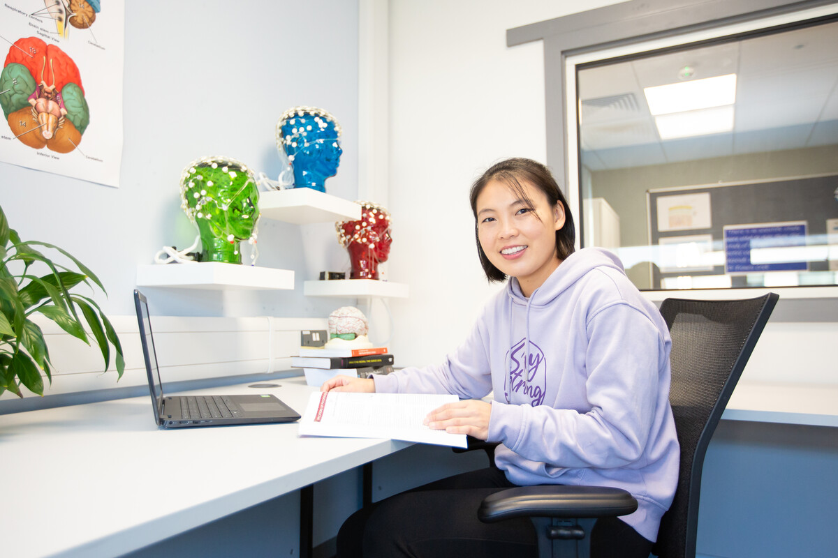 A student is sat at a desk in a Psychology lab, smiling into the camera.