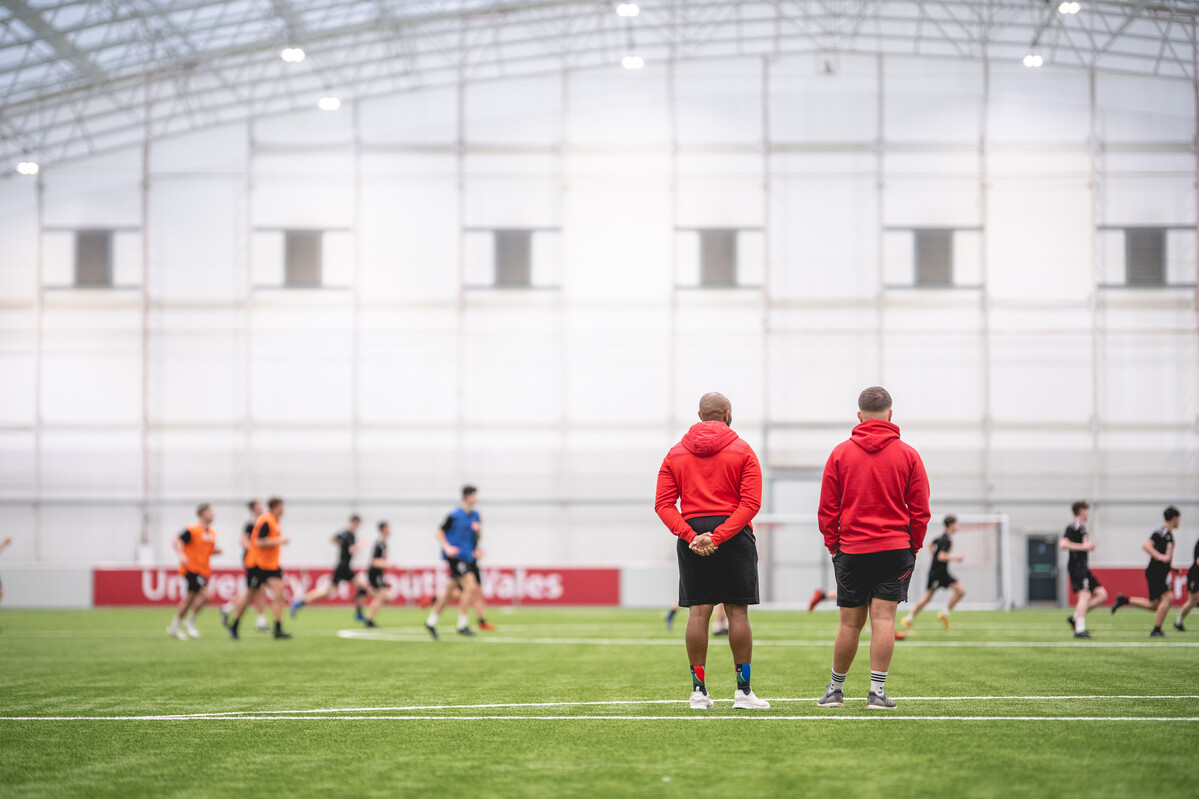 Students playing a game of football on the indoor 3g pitch. Two people are stood on the side line.