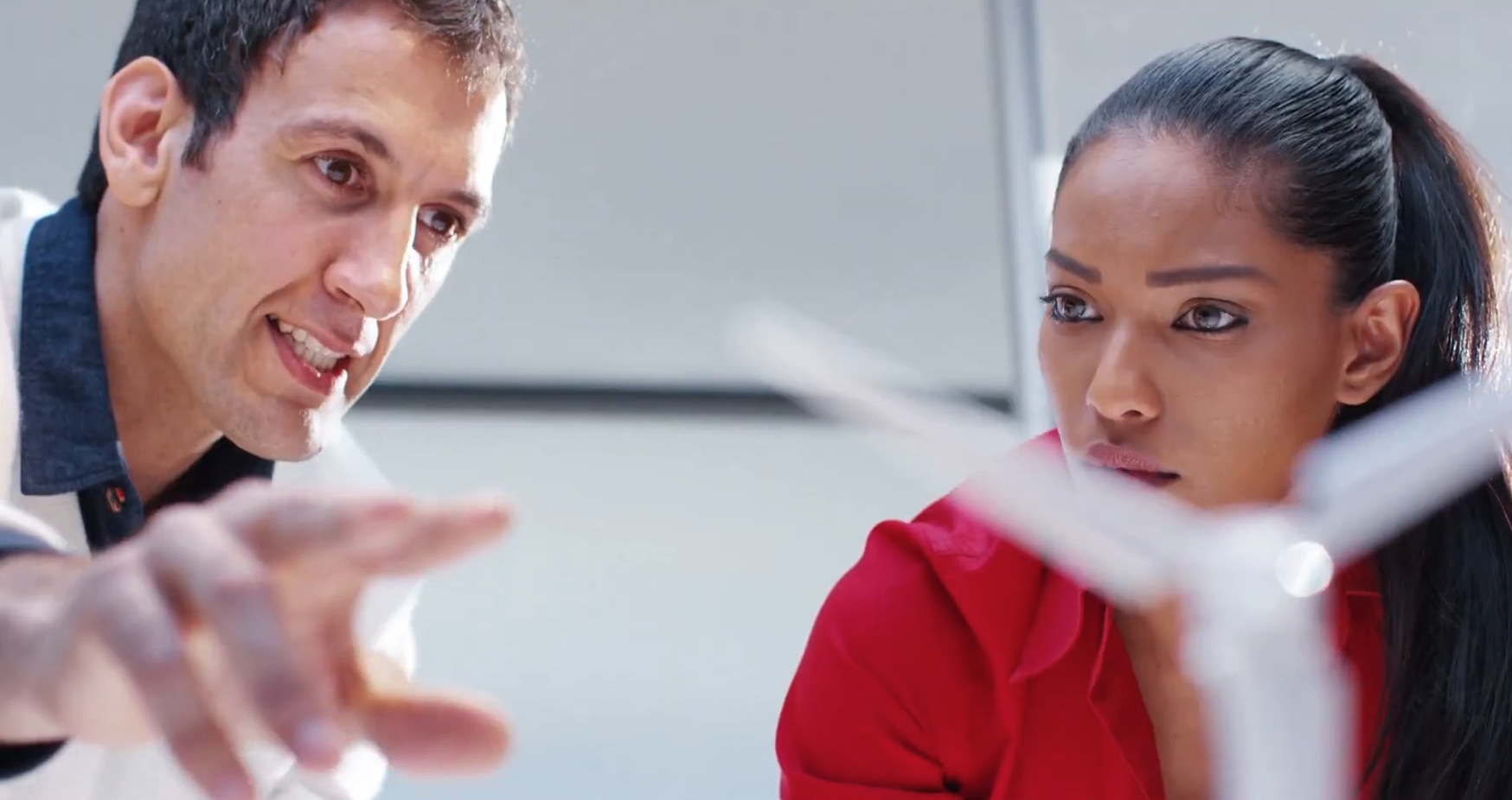 A man in a white sweater and a blue shirt talks with a woman in a red blouse as they look at a miniature wind turbine model