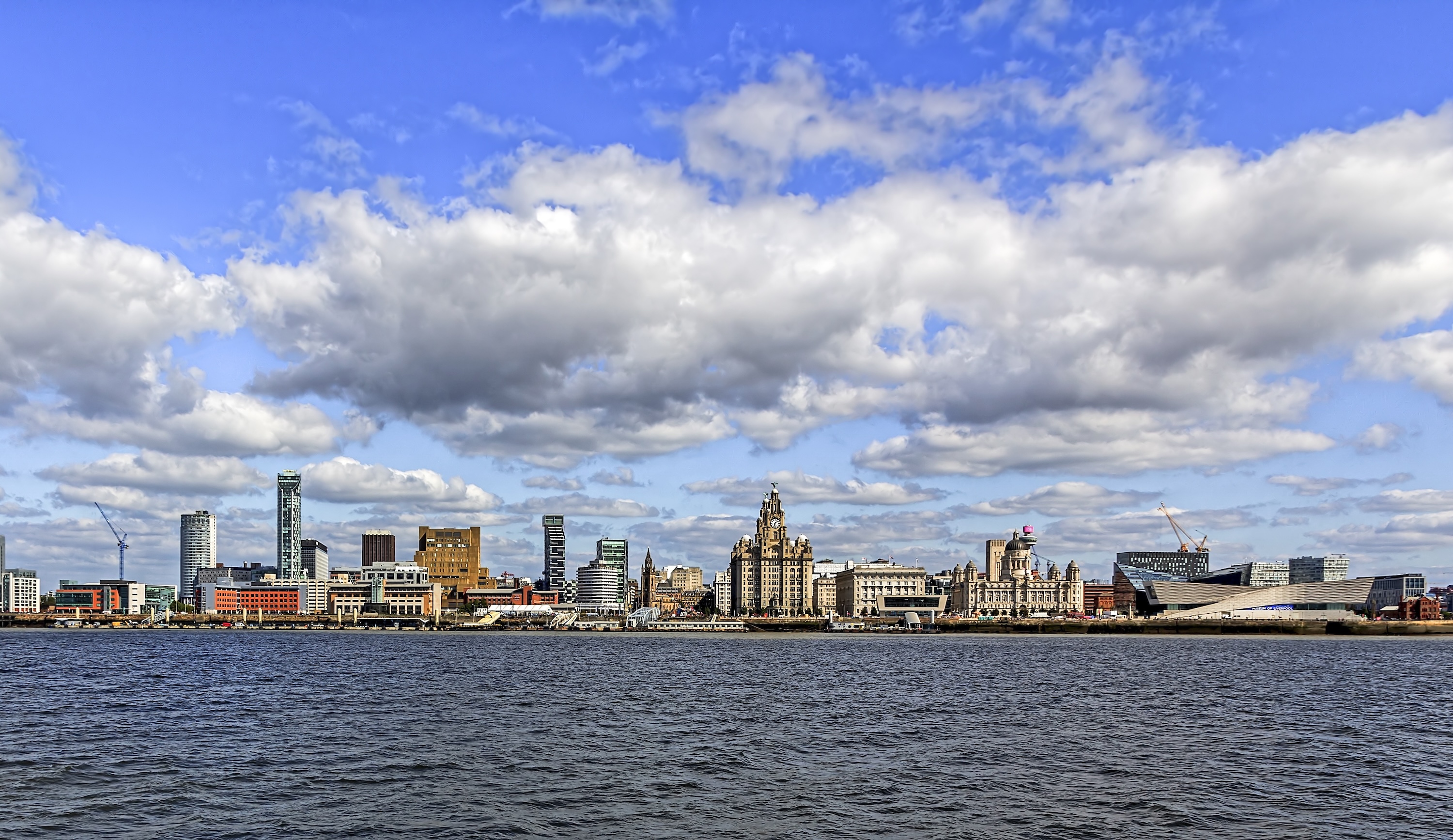 A wide shot of the Cardiff Bay skyline looking at the city from over the water with a blue and cloudy sky in the background