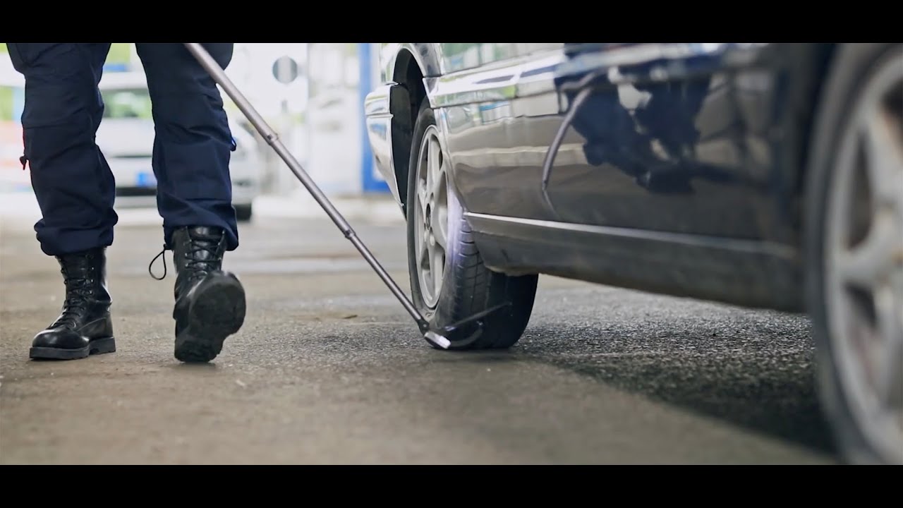 Police officer walking with a mirror on a long pole, looking at the underside of a parked car.