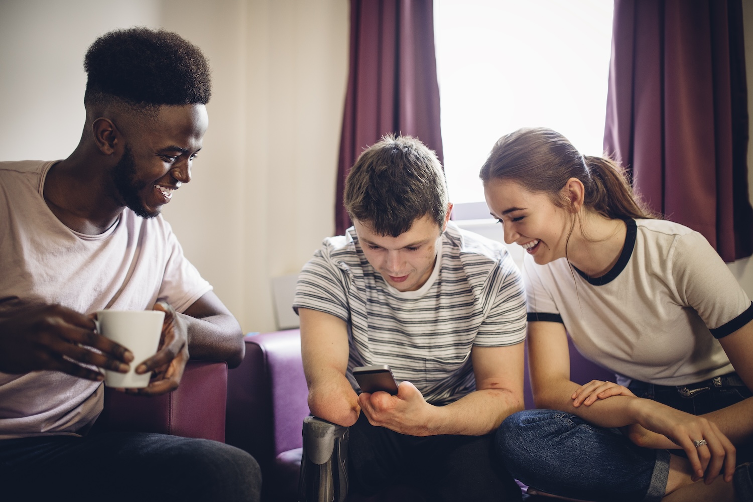 Three diverse friends laugh while looking at a smart phone sat on sofas in student accommodation