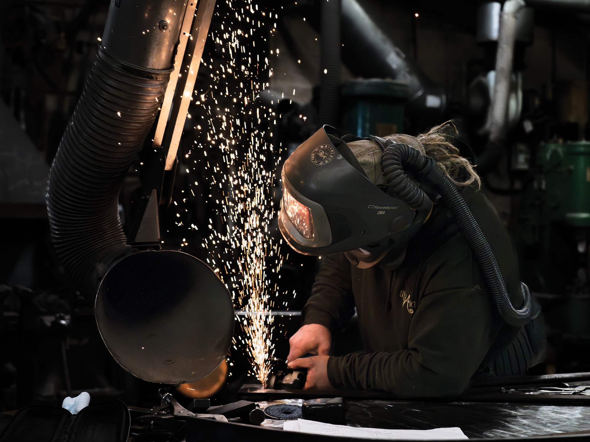 A person in blue overalls and a welding mask welds a piece of metal in a workshop with sparks flying up into the air