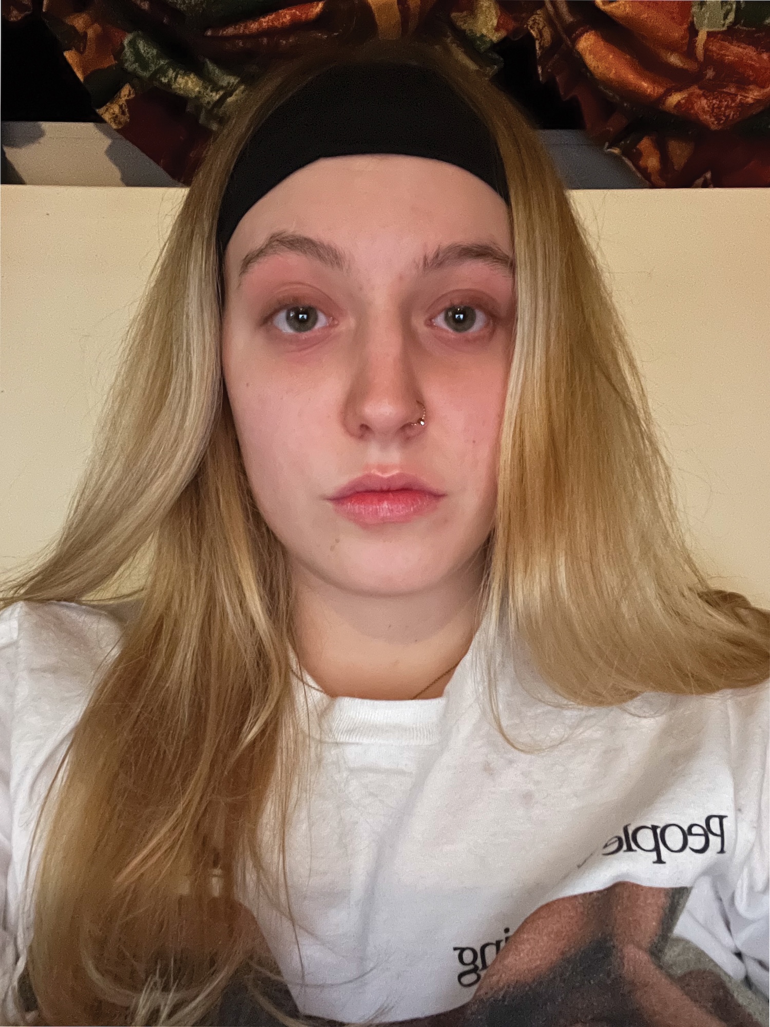 A young woman with blond hair in a white top stairs seriously into the camera with a light yellow wall behind her
