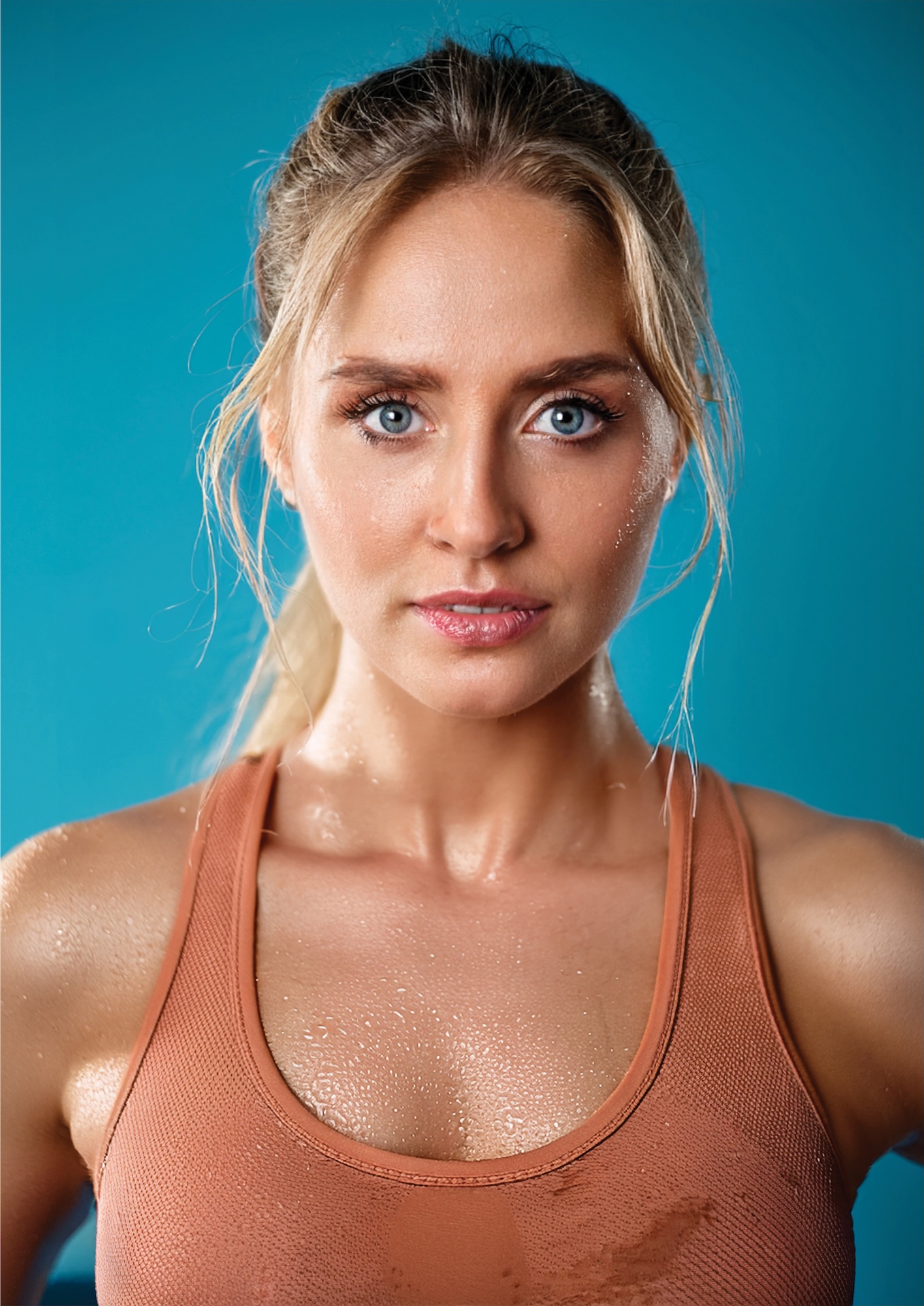 A sweaty-looking young woman in an orange vest stairs intensely at the camera in front of a blue wall