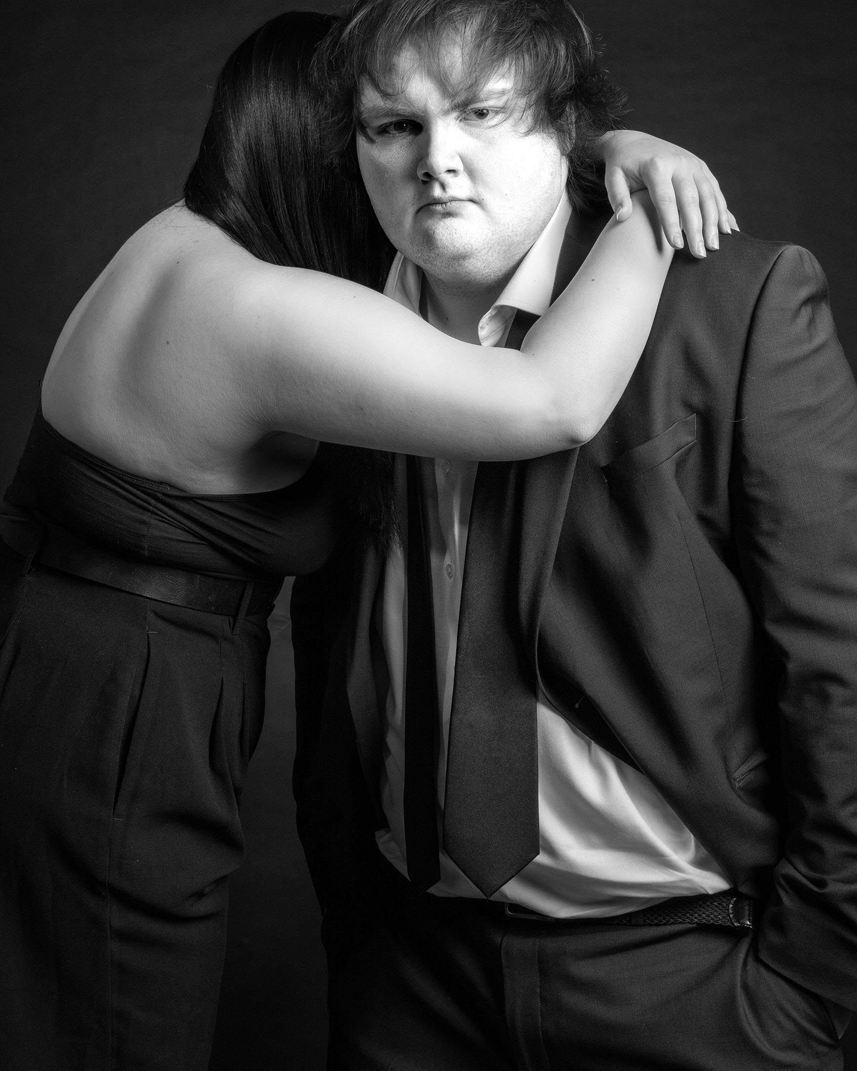 A black and white photo of a woman and a man hugging while the man stairs at the camera