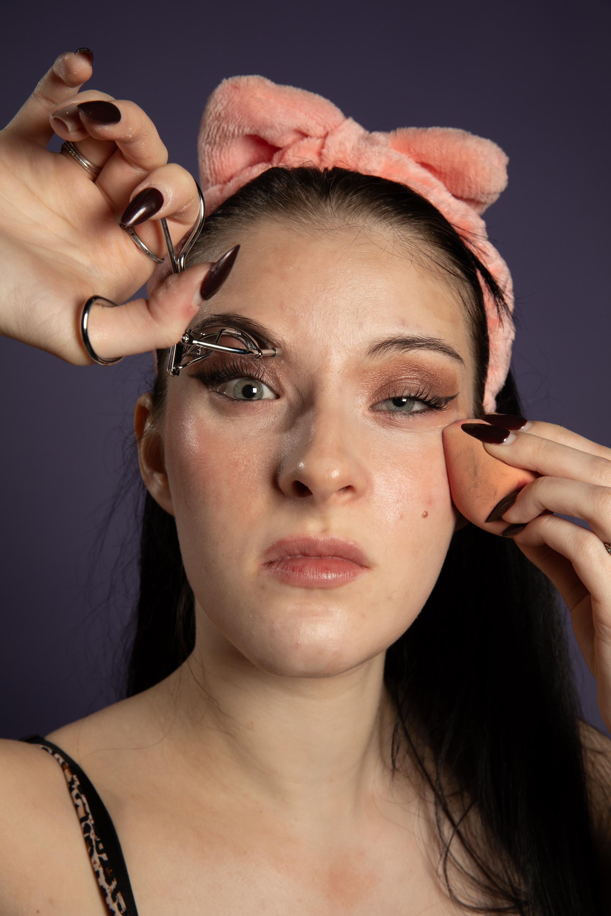 A woman wearing a pink headband uses a make up sponge to apply foundation and uses eye lash curlers while staring intensely at the camera