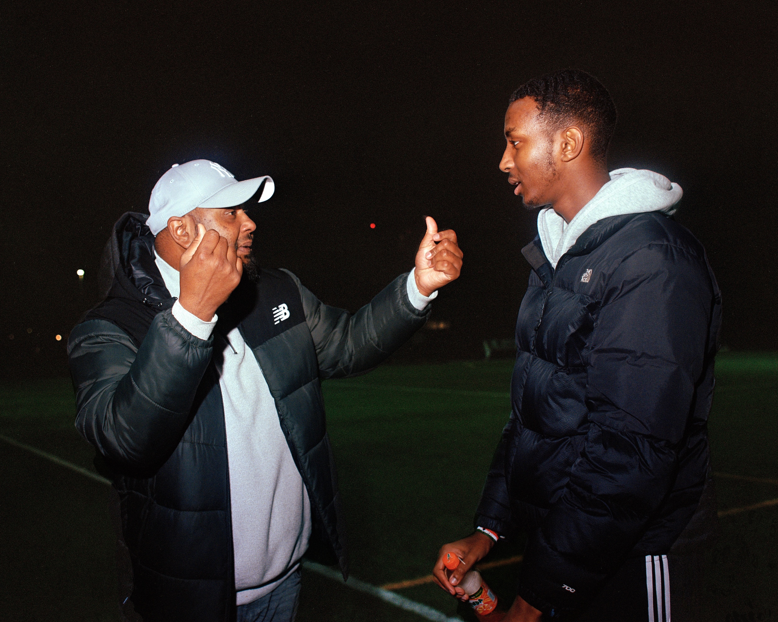 Two men of the global majority and wearing black coats and grey tracksuits talk and gesture with their hands in the dark at a sports field