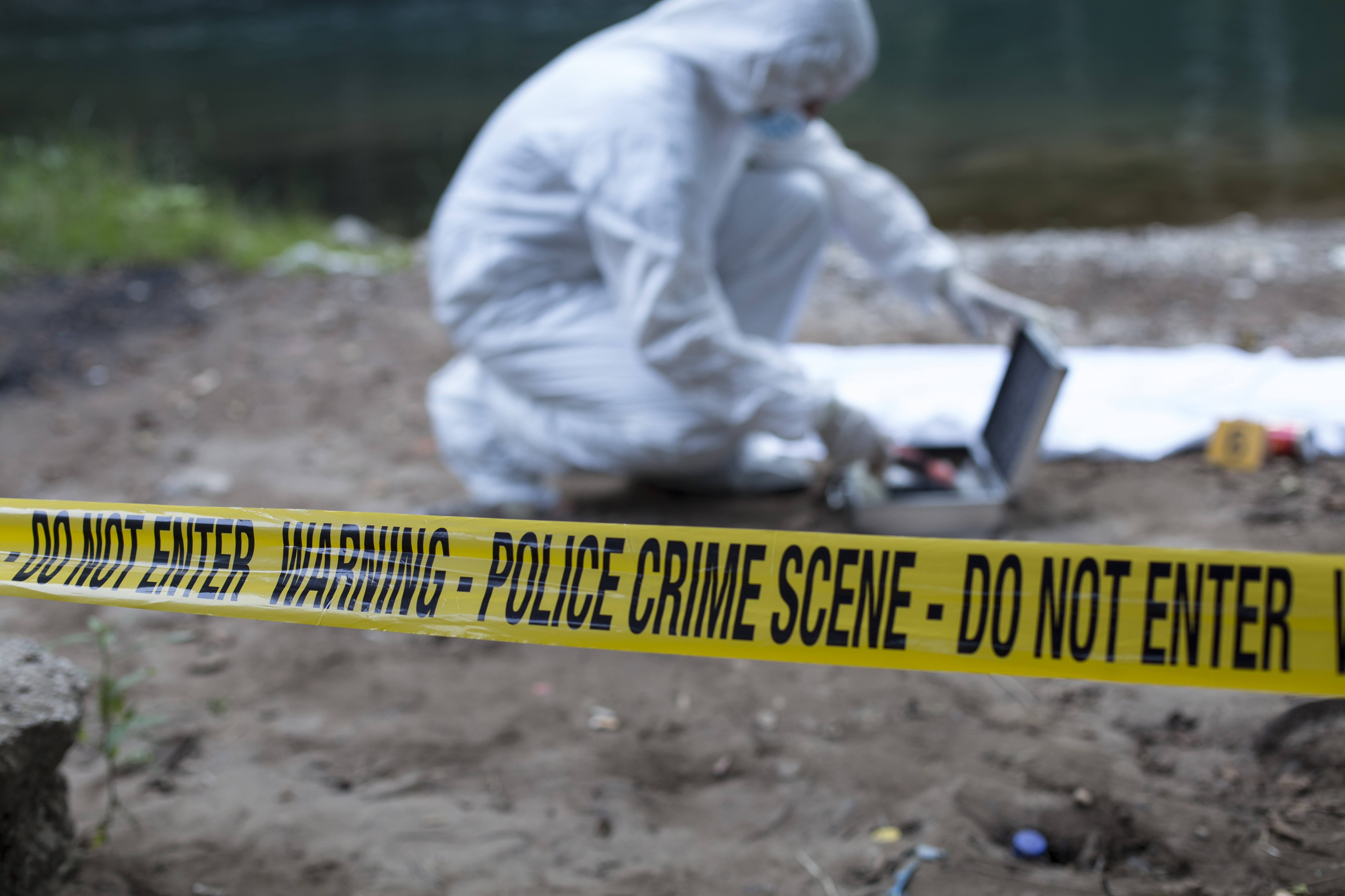 A crime scene investigator dressed in full white overalls looks for tools in a silver case at an active crime scene near a river cordoned off by yellow crime scene tape