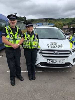 An image of Carwyn and Megan in Police uniform standing in front of a police car.