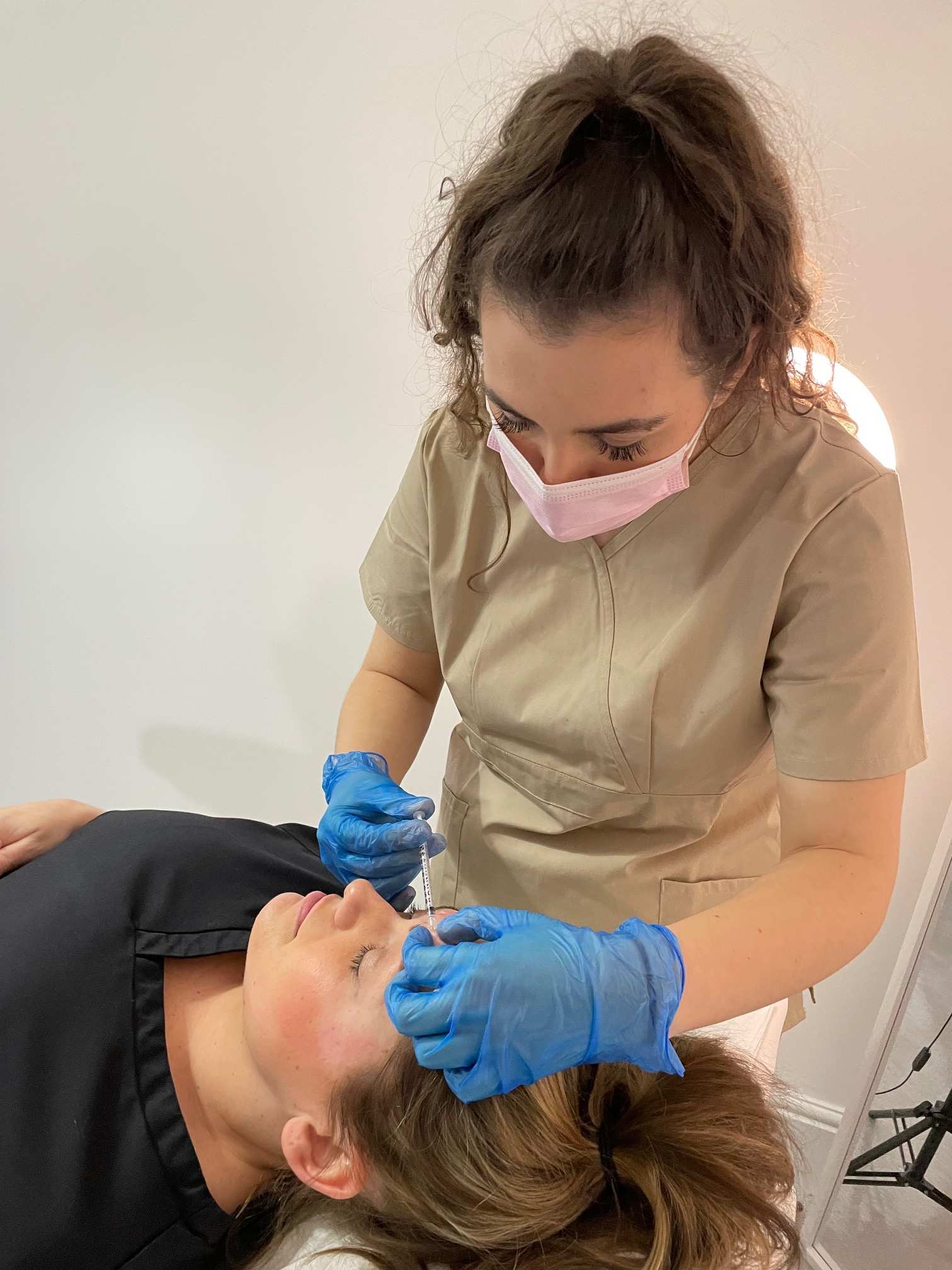 Nursing student Charlotte Jackson-Welford performs a cosmetic procedure on a patient's face with a needle while wearing a white face mask and blue medical gloves
