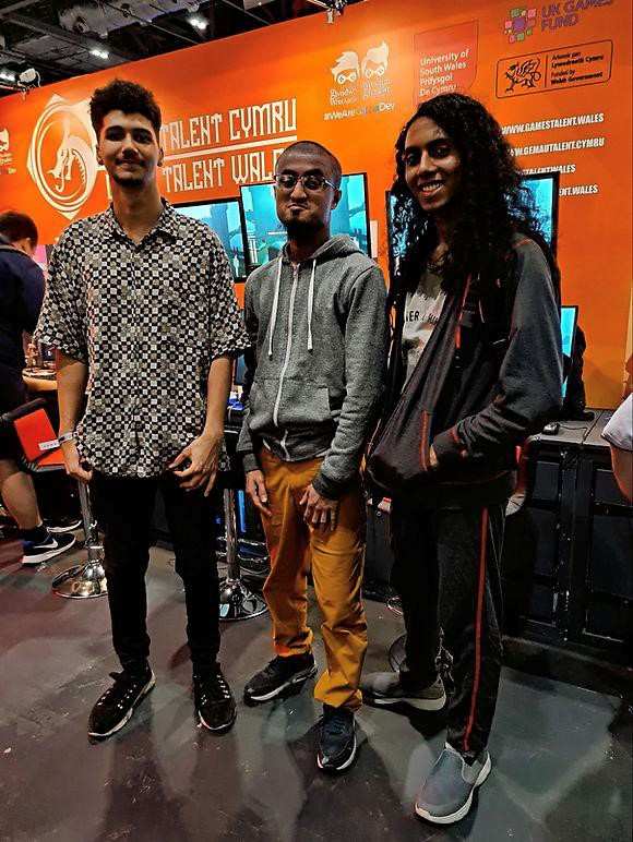 Usman Tarafdar, Jaz Banson, and Kothilmozhiyan Rajavelu stand in front of gaming PCs on a high up desk in front of an orange wall