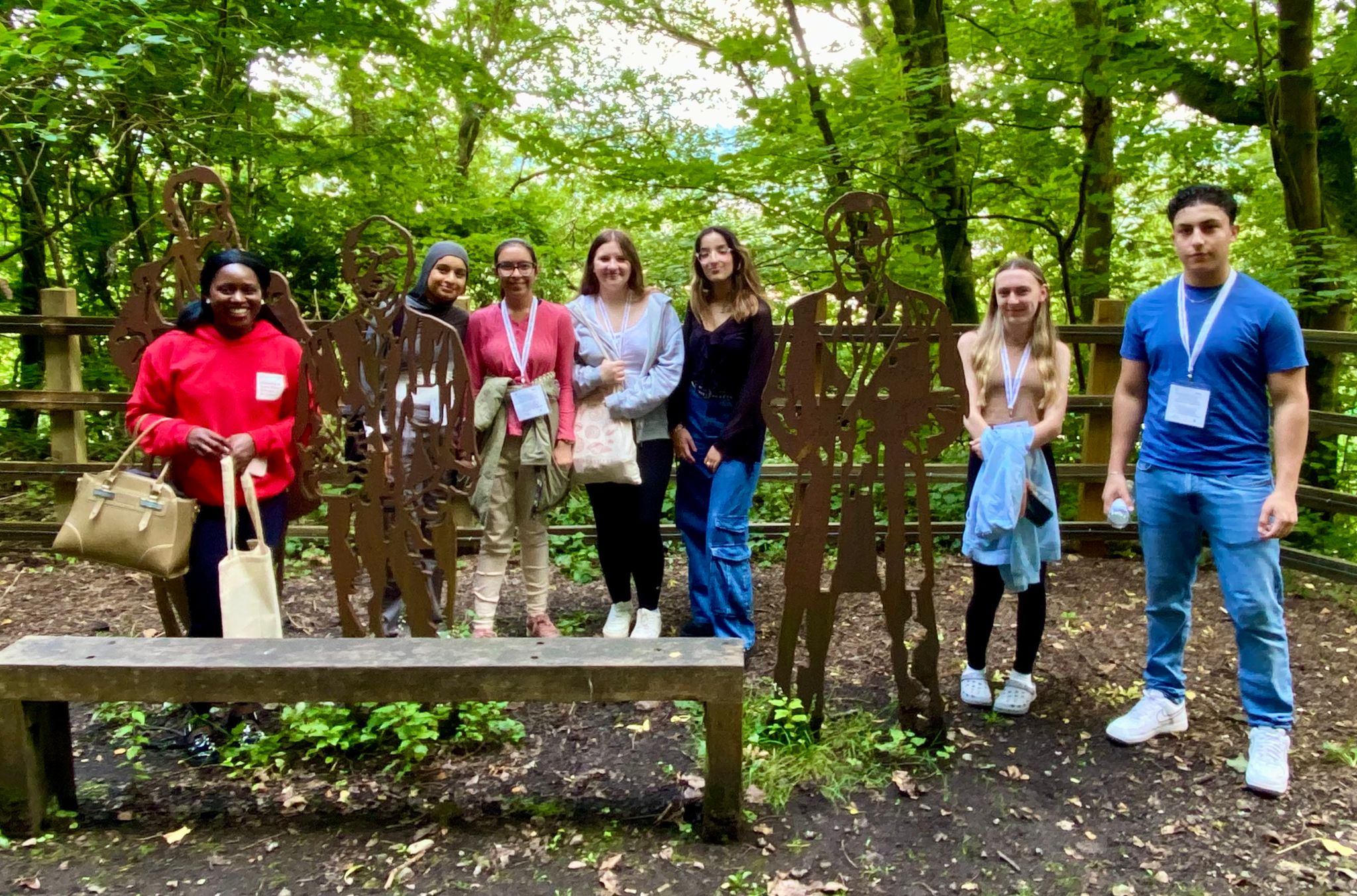 A group of seven people standing outdoors next to metal silhouettes in a wooded area. Some hold bags and wear lanyards, while there is a wooden bench in the foreground.
