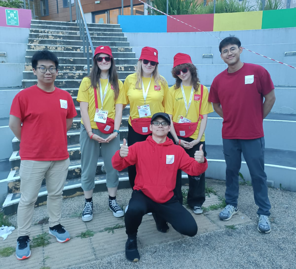A group of five people, four standing and one kneeling, all wearing red and yellow attire, posing for a photo on steps outside. They are all smiling at the camera.