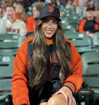 Mae menyw sy’n gwisgo cap du San Francisco Giants a siaced Giants oren a du yn eistedd ar sedd mewn stadiwm yn gwylio gêm bêl fas.