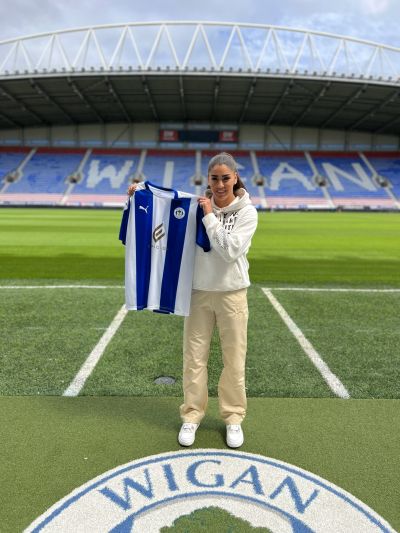 Mae person yn sefyll ar gae pêl-droed yn dal crys Wigan Athletic glas a gwyn. Mae seddi stadiwm a'r llythrennau mawr 