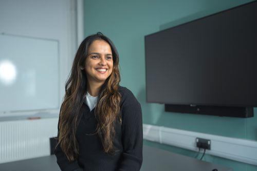 Chamathka is stood in a classroom in front of a large TV screen. She is wearing a black jumper and smiling for the camera.