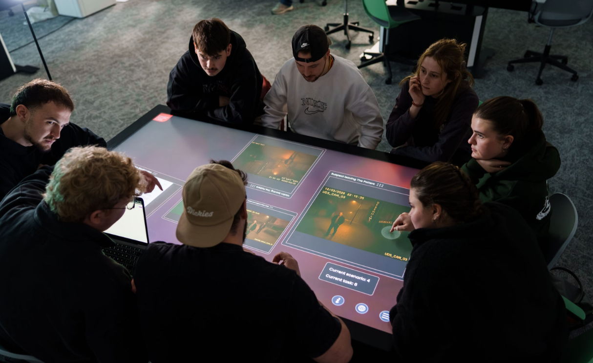 A group of eight students gathered around a large interactive touchscreen table, collaborating on a digital project. Several are pointing at the screen, which displays multiple image panels and interface controls, while others watch and discuss in a modern classroom or lab setting.