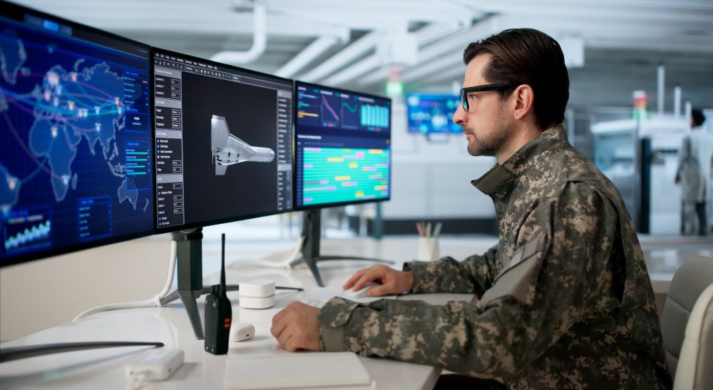A person in military uniform sits at a workstation monitoring multiple computer screens displaying maps, data, and graphics in a command and control environment.